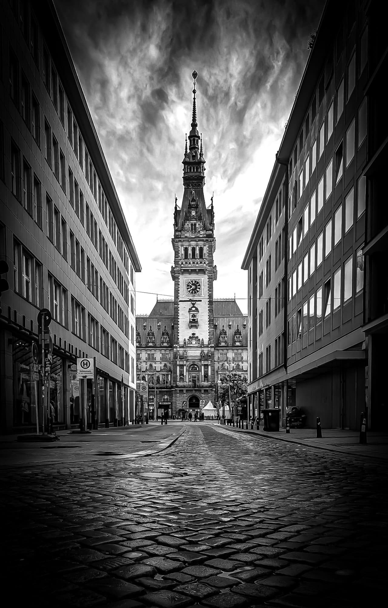 Black-and-white street view framed by tall modern buildings on both sides, leading along cobblestones to a large historic clock-tower building centered in the distance under a cloudy sky.