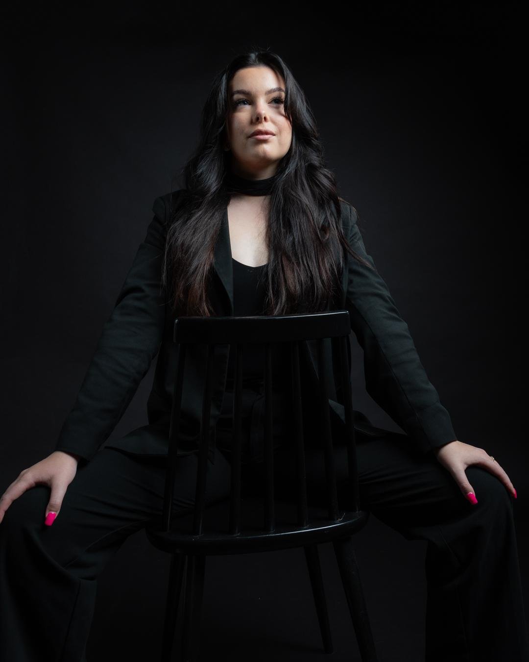 The image shows a person dressed in black sitting on a chair, posed in a confident manner. The background is dark, which puts the focus on the person in the image. Valokuvaaja Lappeenranta Kuvaajankulma
