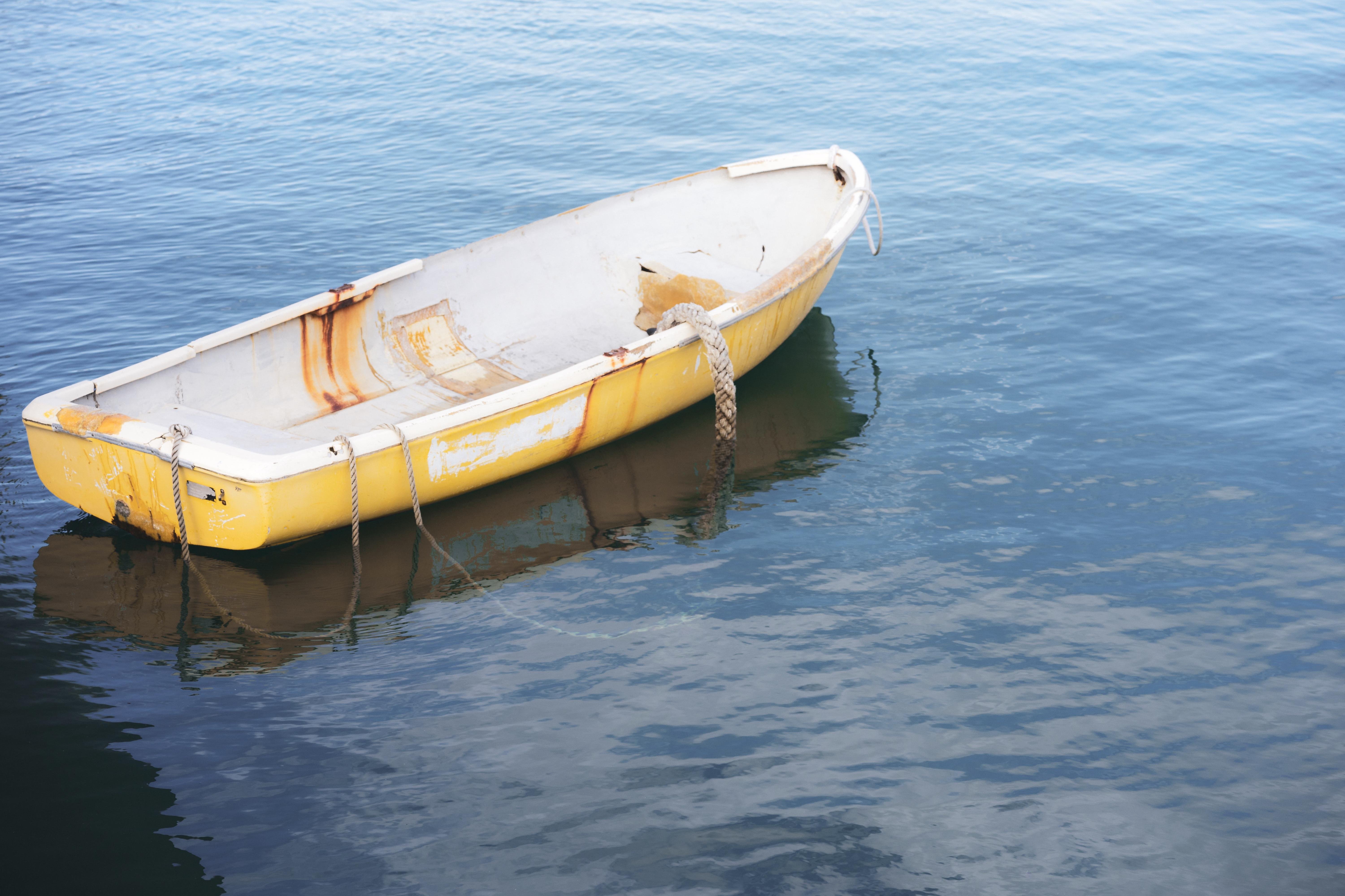 The image shows a small yellow rowboat floating on calm water. The boat has ropes attached, and there are visible signs of rust and wear on its surface. The surrounding water is blue and reflects the boat's color.