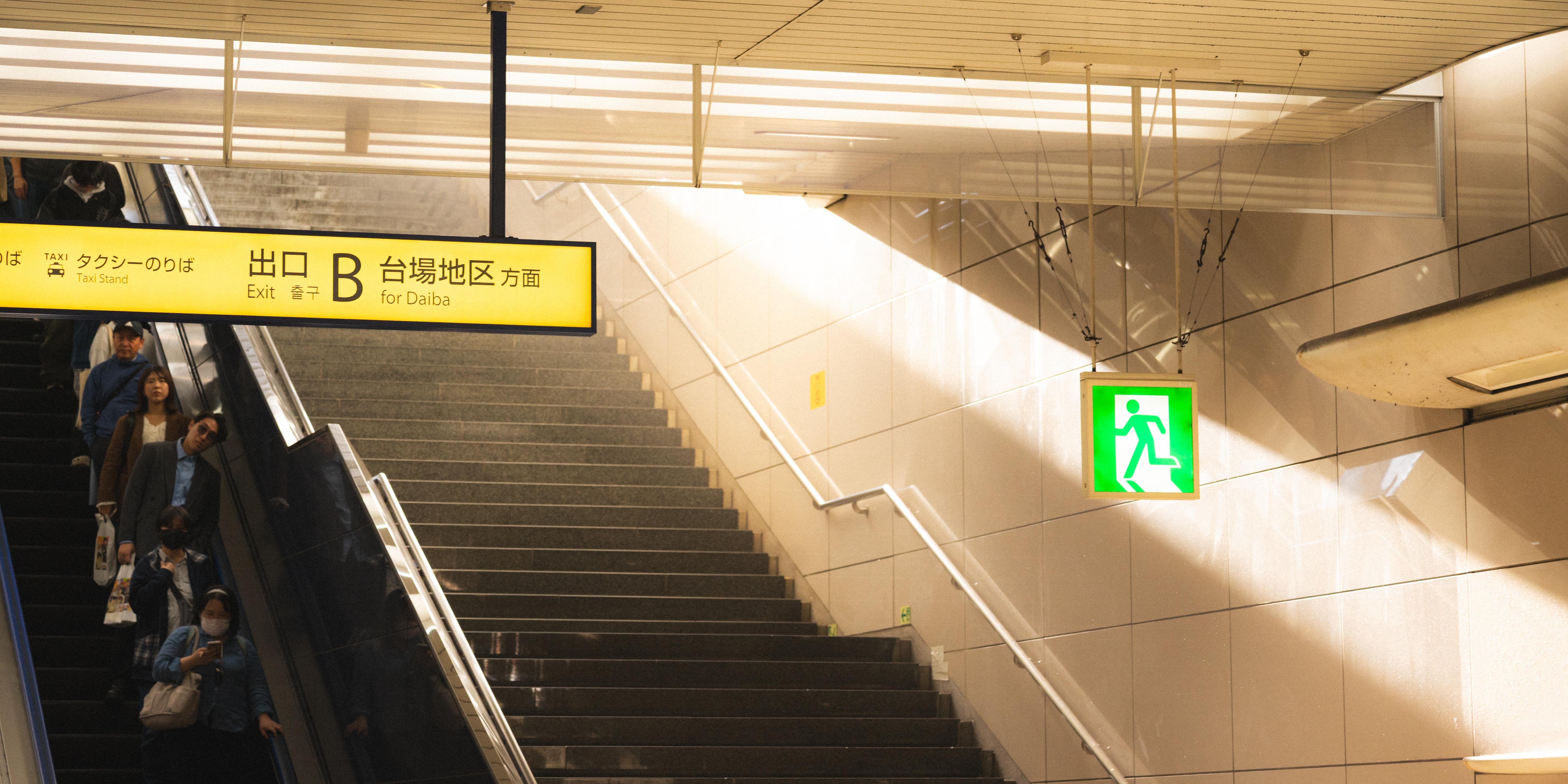 People descending an escalator alongside a staircase in a subway station. A yellow sign overhead indicates Exit B for Daiba. A green emergency exit sign is visible on the wall. Bright overhead lighting creates reflections on the tiled surfaces.
