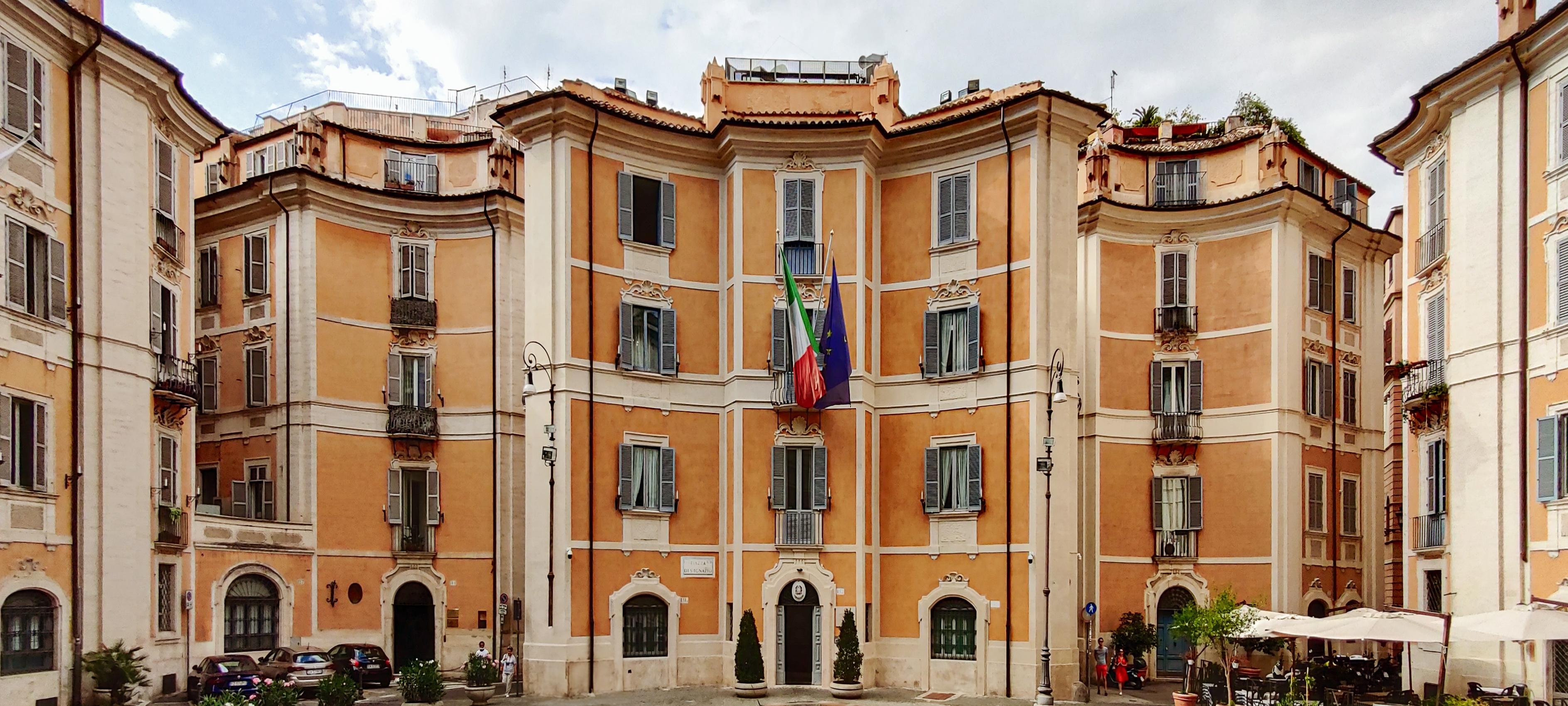 Un edificio con una facciata curva in stile architettonico classico. Presenta numerose finestre con persiane e due bandiere sono esposte sopra l'ingresso. L'area circostante include alberi, piccoli tavoli e sedie, suggerendo la possibilità di un caffè all'aperto o un'area piazza.