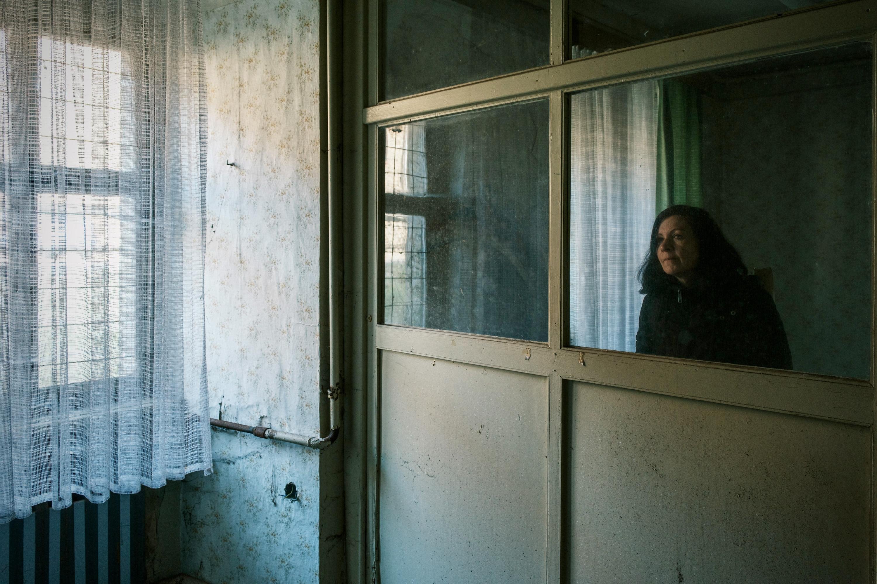 A woman with dark hair sits in a room, gazing at her reflection in a dusty glass partition. The walls are decorated with floral wallpaper, and sheer curtains filter natural light from a nearby window. The overall atmosphere feels nostalgic and contemplative.