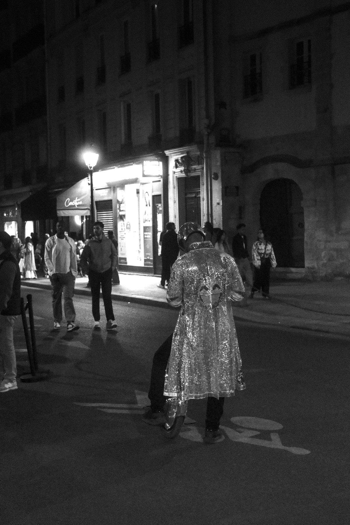 Photo verticale en noir et blanc. Dans la rue, un homme de dos avec un manteau trenchcoat couvert de facettes réfléchissantes comme une boule disco, un pied à terre, l’autre sur un monocycle électrique, tête casquée style disco aussi, entrain de regarder son téléphone