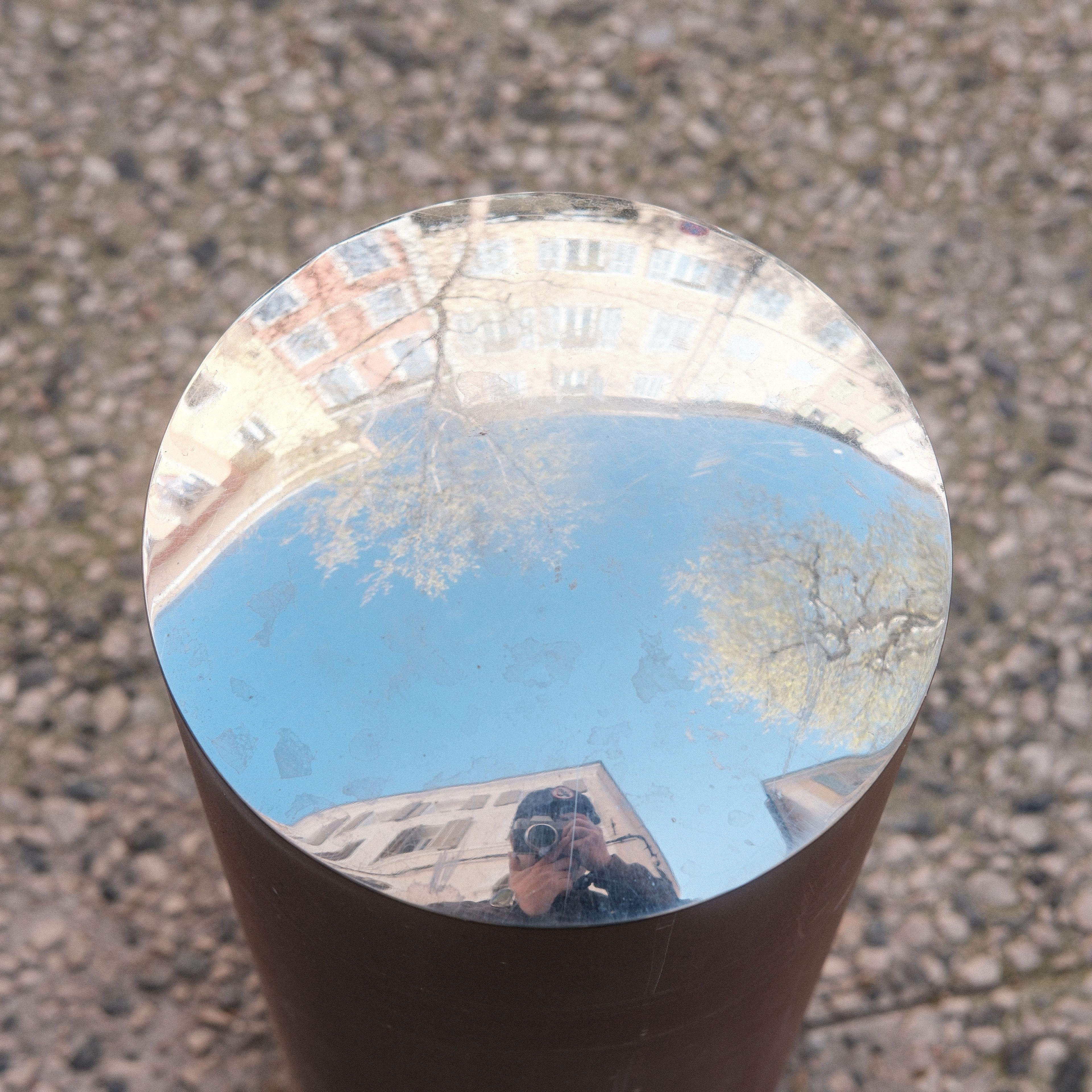 Photo carrée. Le haut d’un petit poteau de rue dont le sommet est en métal faisant un miroir concave. On y voit le reflet de petit immeubles de 3 étages, le ciel bleu, des arbres en fleur et le photographe, l’appareil photo devant son visage