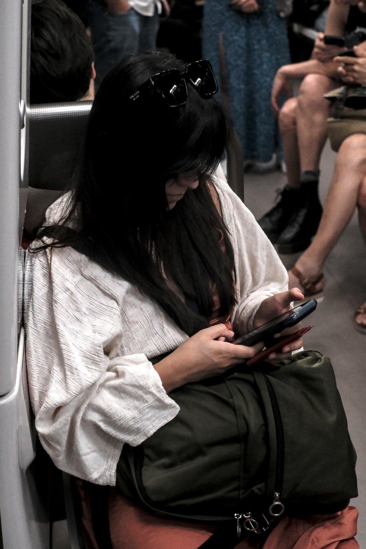 Photo verticale. Une femme au cheveux long noir lâchés, lunettes de soleil posés sur la tête, une frange lui cachant le visage, un haut ample blanc, assise sur un strapontin d’un métro, entrain de consulter son téléphone