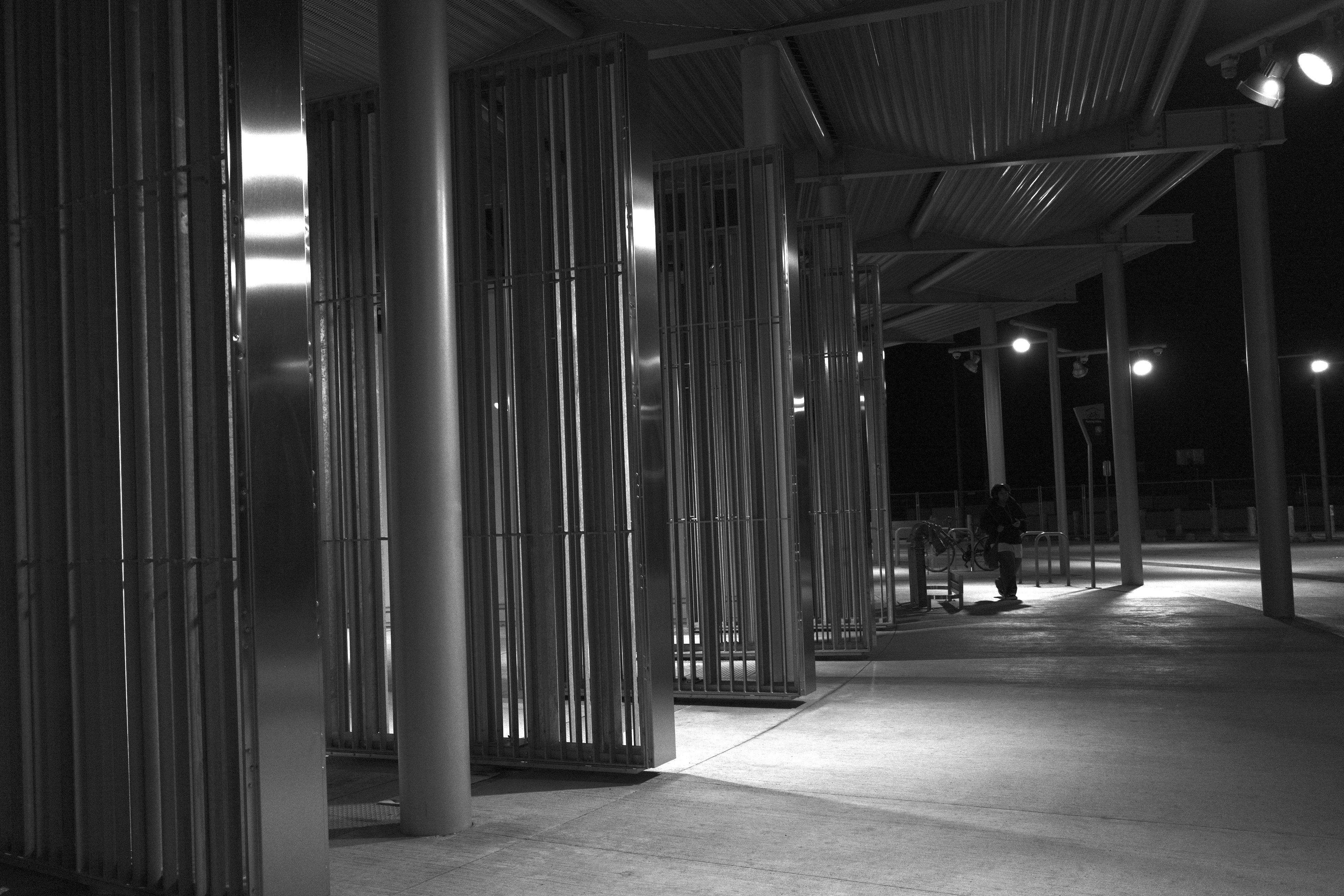 Photo horizontale en noir et blanc, de nuit. S’étends depuis la gauche en arc de cercle, des grands portaux à barreaux, ouverts. De la lumière émane du bâtiment à gauche