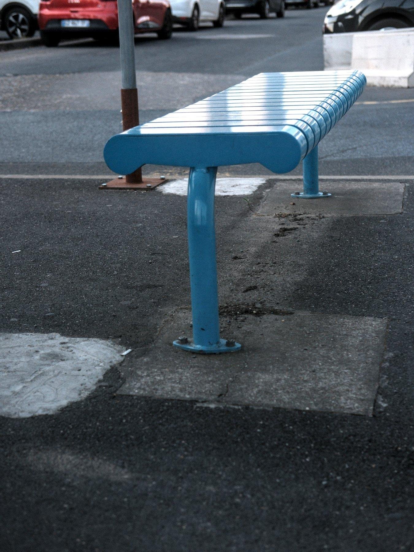 Photo horizontale. Un banc bleu fixé sur un sol en bitume. On devine un parking derrière