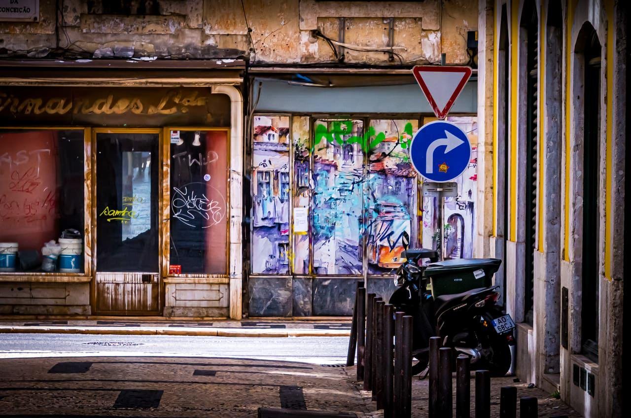 Narrow urban street framed by aged buildings with faded paint and graffiti. A closed storefront displays graffiti on its doors, while a blue and white traffic sign indicates a right turn. A motorcycle is parked near the buildings on the right side. The street surface is composed of cobblestones with sunlight illuminating the scene.