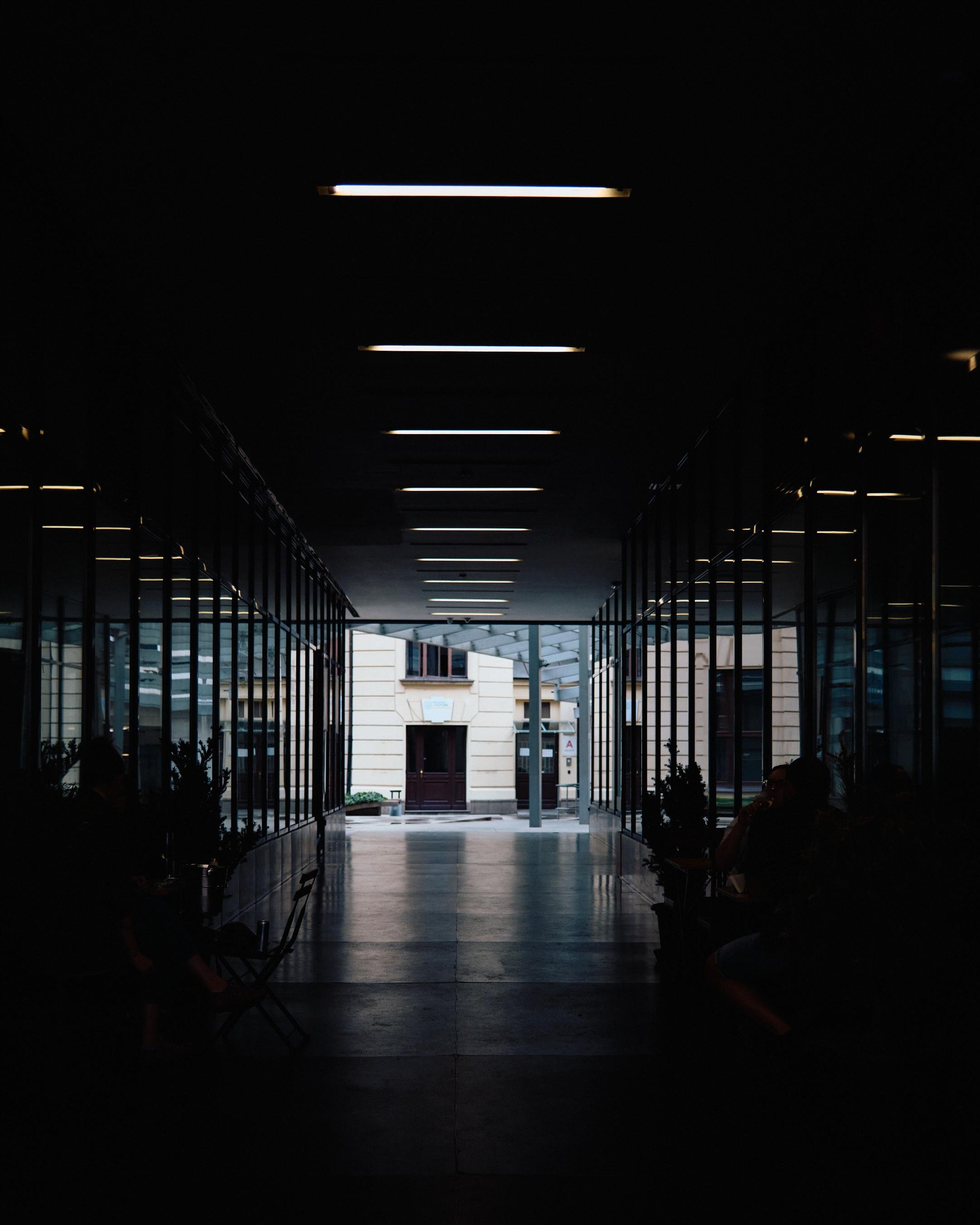 Long, symmetrical corridor with dark ceiling and evenly spaced rectangular lights, glass walls on both sides, and a reflective floor leading to an outdoor doorway and building facade at the far end.