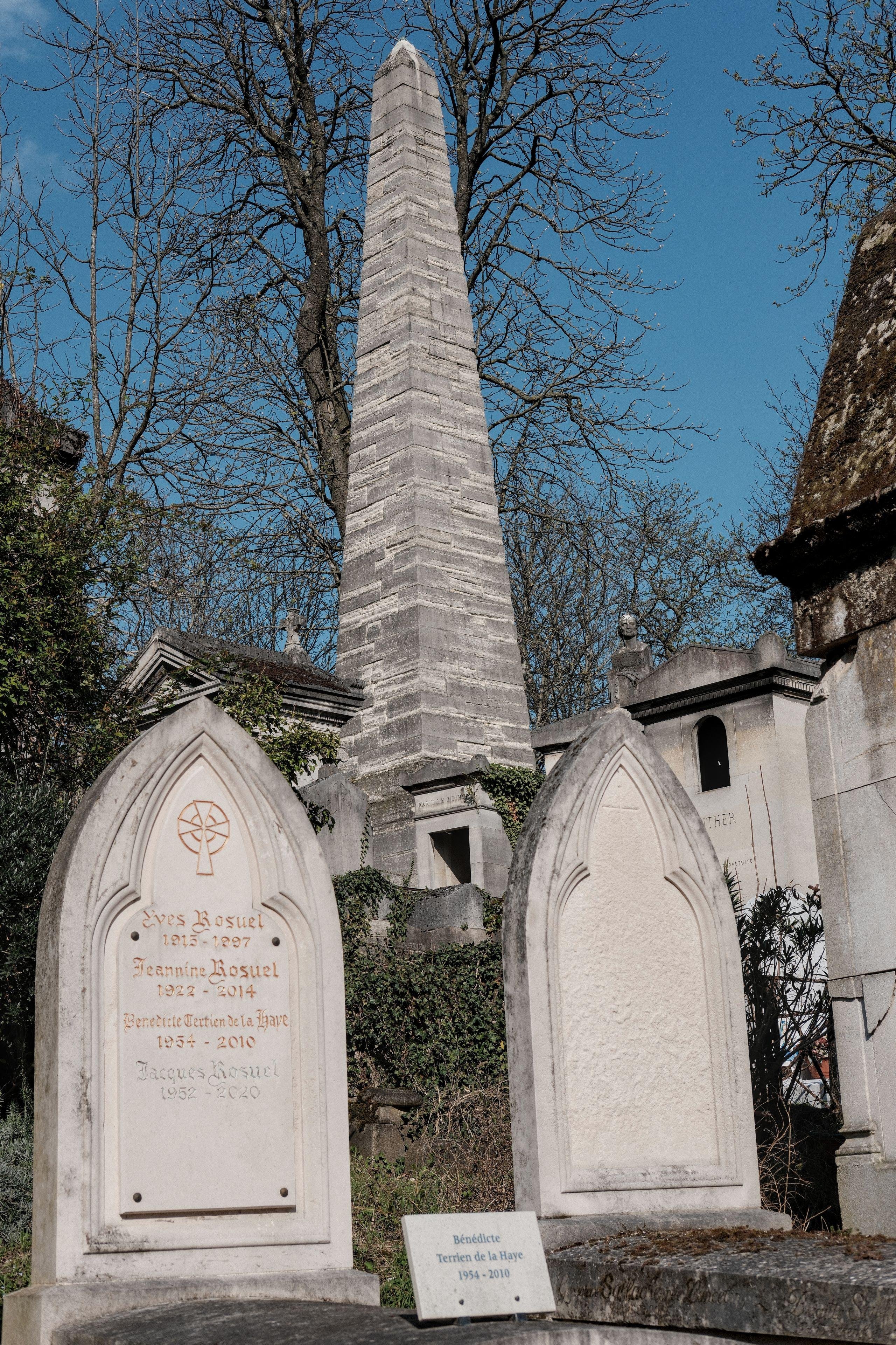 Photo verticale. Au premier plan, deux pierres tombales avec le haut pointu et derrière, une autre tombe avec une tombe surmontée d’un obélisque en pierre. Le tout sur fond de branches d’arbres et de ciel bleu