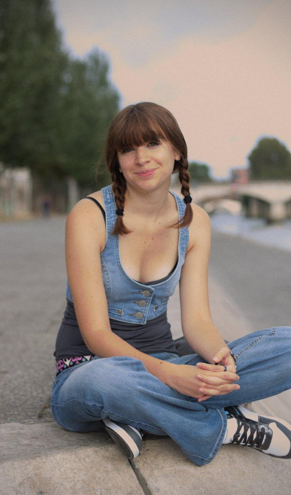 Une fille brune assise en tailleur sur les quai de la Seine. Elle est en jean et avec un débardeur bleu très sombre, avec par dessus un gilet sans manche en denim aussi. Elle a deux nattes de part et d’autre du visage. Elle regarde la caméra en souriant.