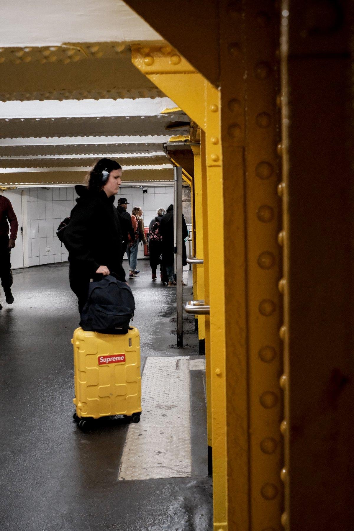 Photo verticale. Sur la droite, alignement de pylône en acier rivetés peints en jaune Ligne 1 du métro parisien. Sur leur gauche, un homme habillé en noir avec des un casque audio sur la tête fait rouler à sa droite une valise du même jaune avec posé dessus un sac à dos noir.