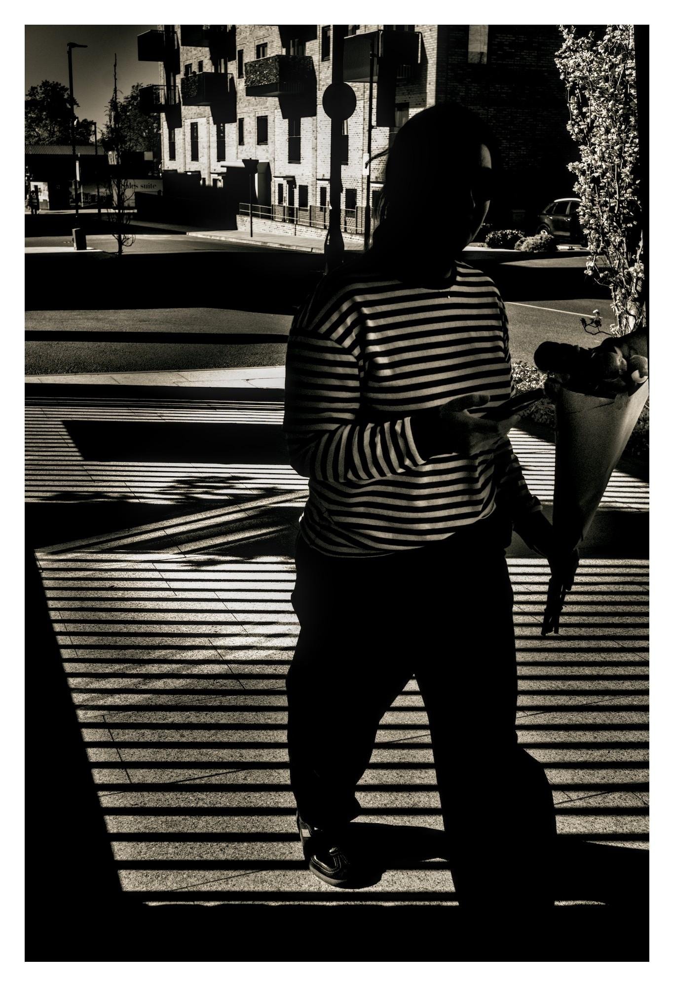 Silhouetted person in a striped long-sleeve shirt standing on a sunlit sidewalk with strong horizontal shadow lines, holding a cone-shaped bouquet; apartment buildings, street signs, and parked cars in the background.