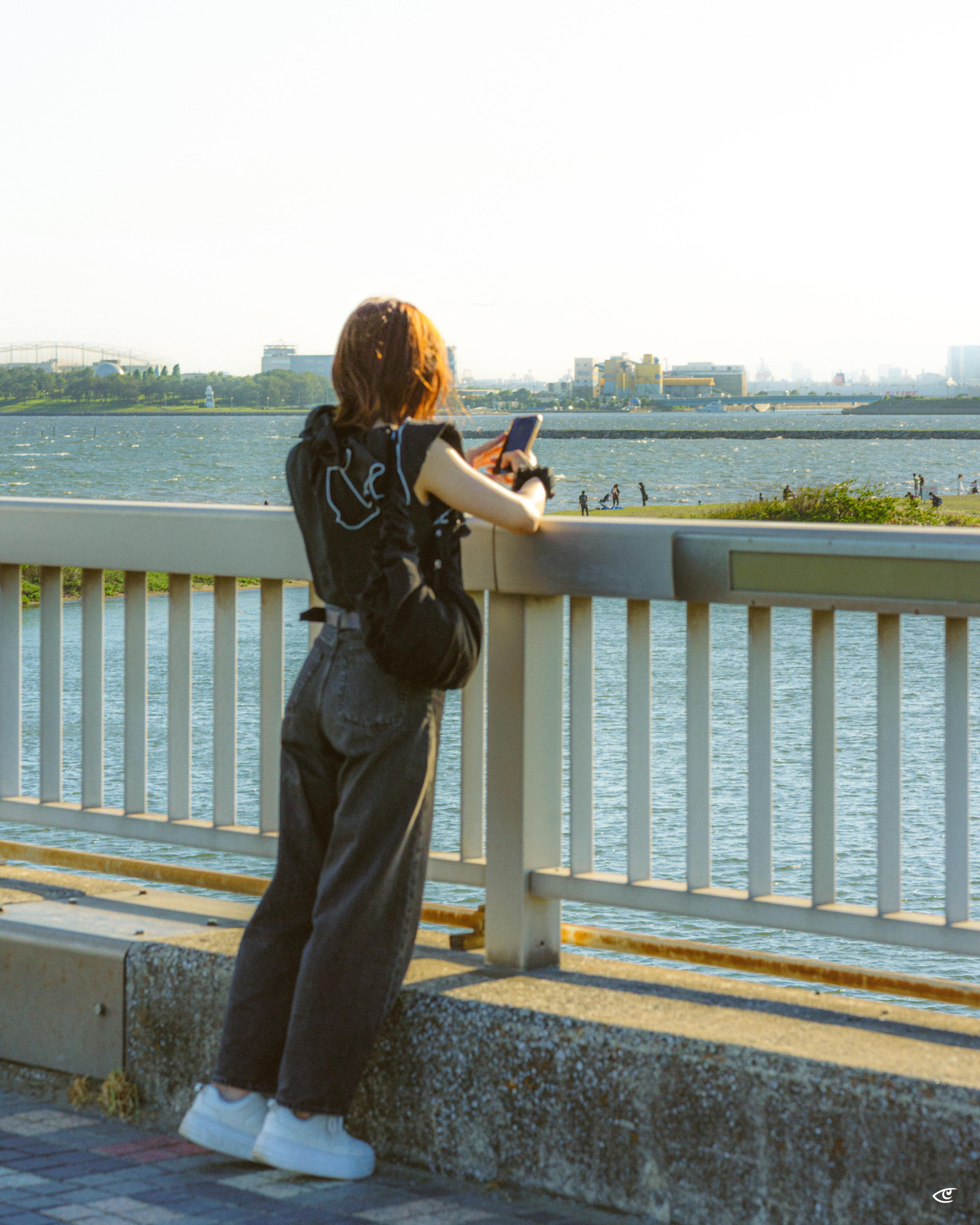 Person with short brown hair wearing a black backpack, dark pants, and white shoes leans on a white railing while looking at a smartphone, with a wide river and distant buildings in the background.