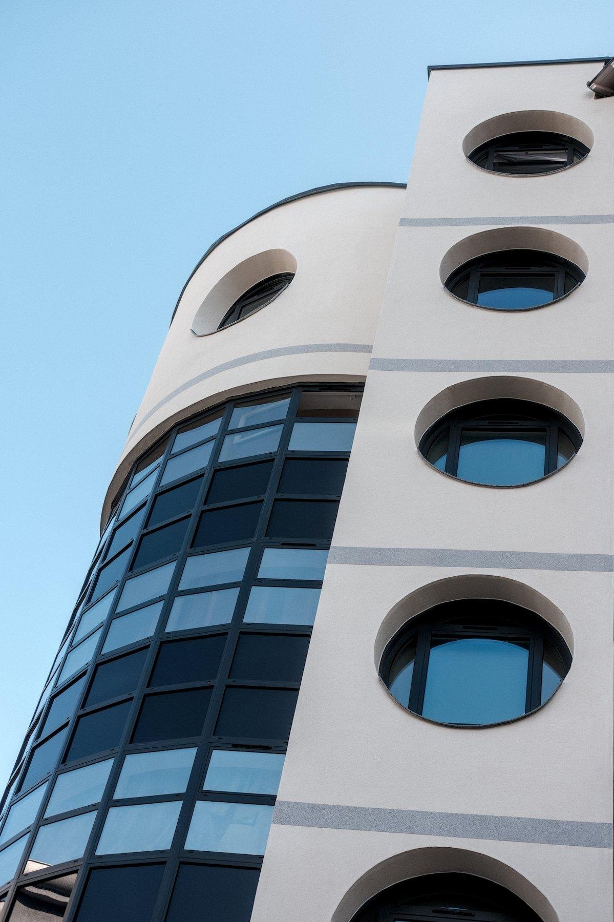 Photo verticale. Contre-plongée sur un bout de bâtiment moderne, blanc, avec des fenêtres rondes à chaque étage, et sur la gauche, un décrochage cylindrique en baie vitrée sur toute la hauteur sauf au niveau du dernier étage, ou une fenêtre ronde est visible et laisse supposer l’existence d’une autre de l’autre côté du cylindre. Le tout sous un ciel bleu se réfléchissant aussi dans le verre
