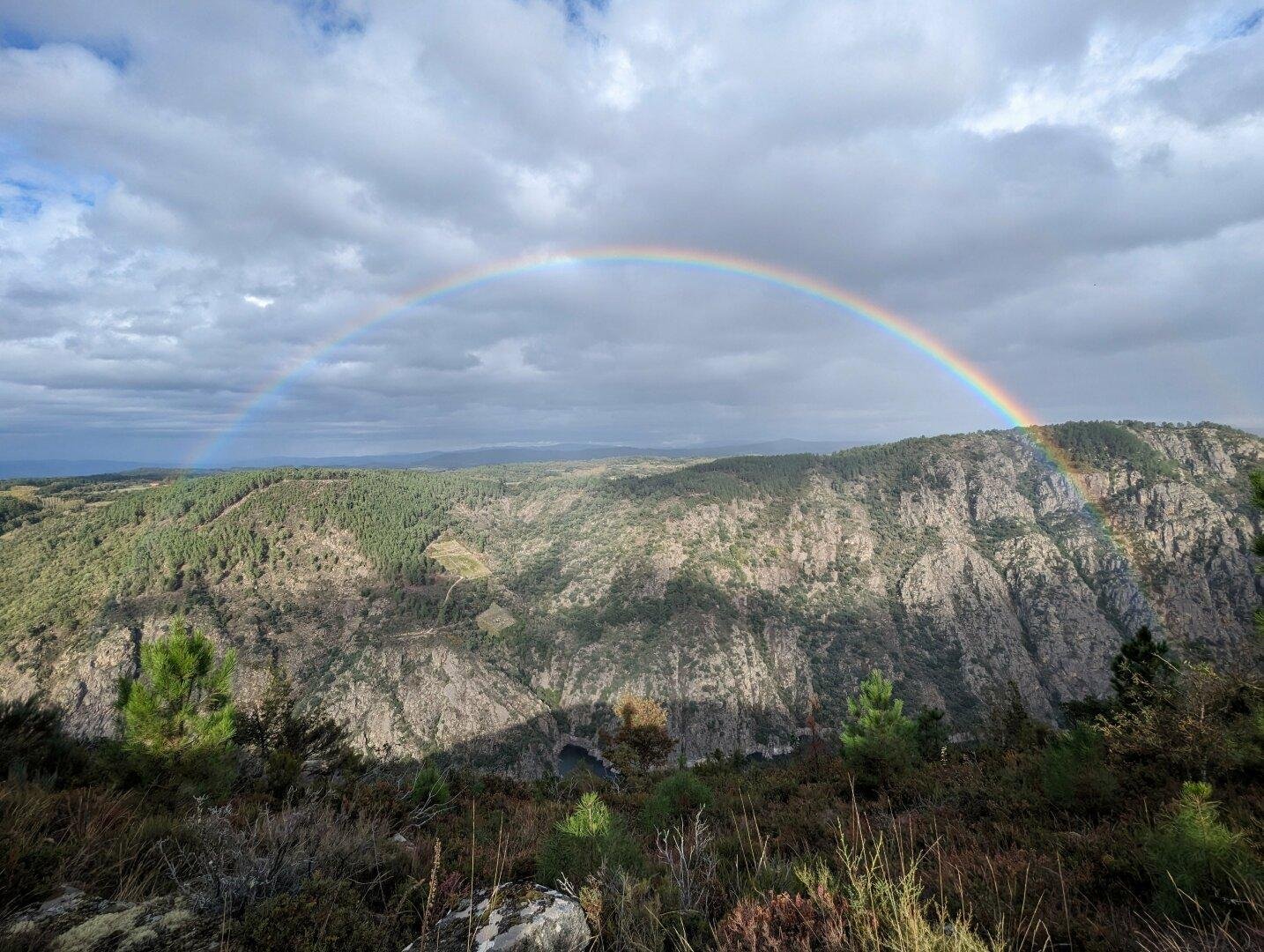 Paisaje montañoso. Se ve una montaña subiendo desde una gran depresión, un cañón erosionado en la piedra. Ya más al fondo, un inmenso arco iris sirve de ribete a toda la montaña que se eleva sobre el cañón. De derecha a izquierda de la foto se ve el arco iris nítidamente atravesando el paisaje. El cielo tiene unas nubes algo grises, con algún claro ocasional. El sol baña parte de la ladera de la montaña, haciendo brillar la piedra, de color claro. Parece un paisaje idílico, de sueño.
