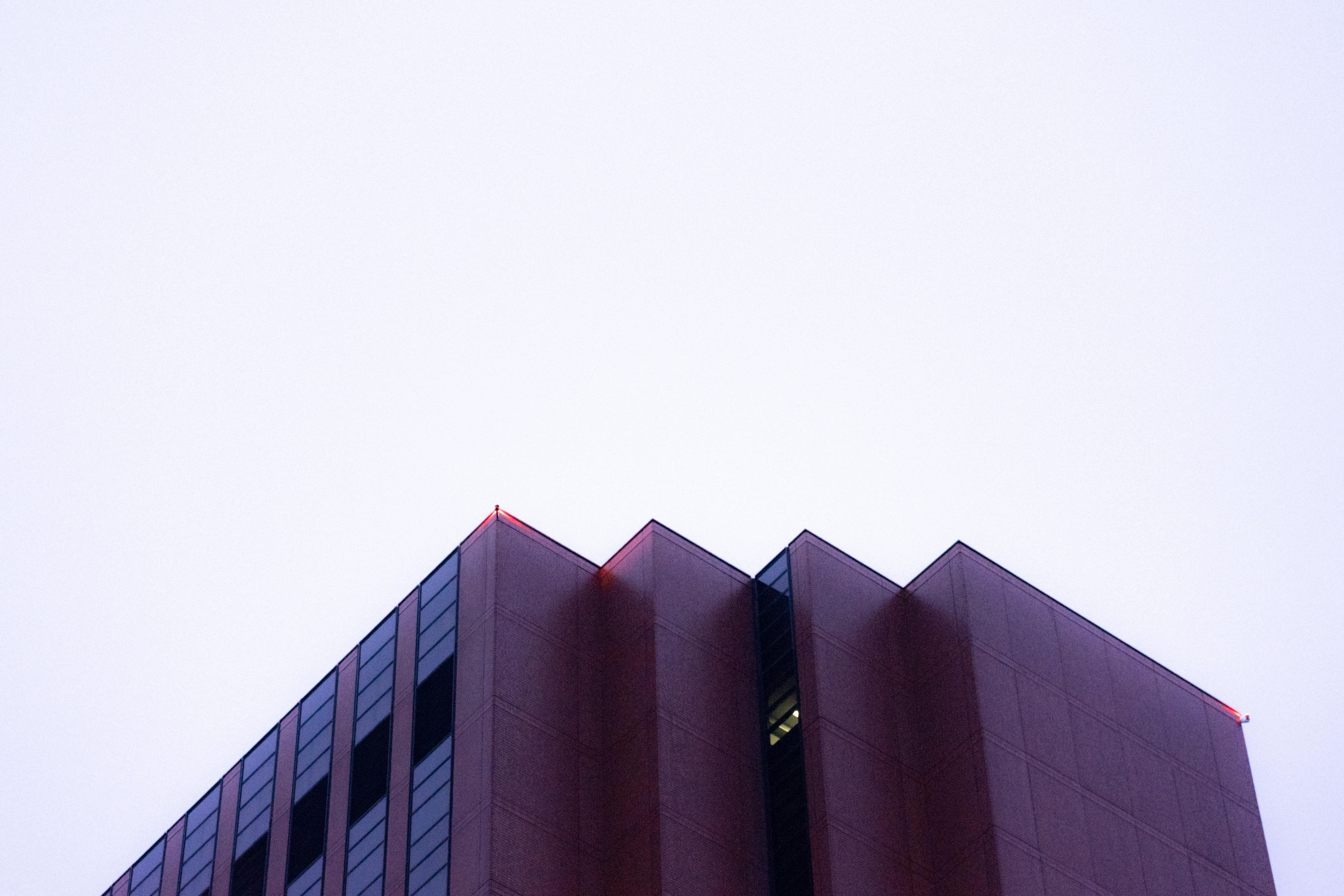 Top corner of a tall, modern building with a sleek facade, featuring a combination of windows and solid panels. The sky is overcast, creating a stark contrast with the building's dark exterior. The edges of the roof have small red lights. One lit window is visible near the center of the building.