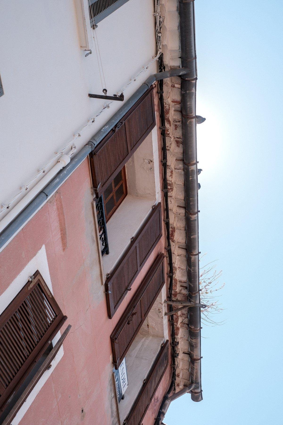 Photo verticale. Contre plongée sur la gouttière d’une maison rouge pastel où on voit presque à contre jour, 3 pigeons avec le halo solaire derrière eux