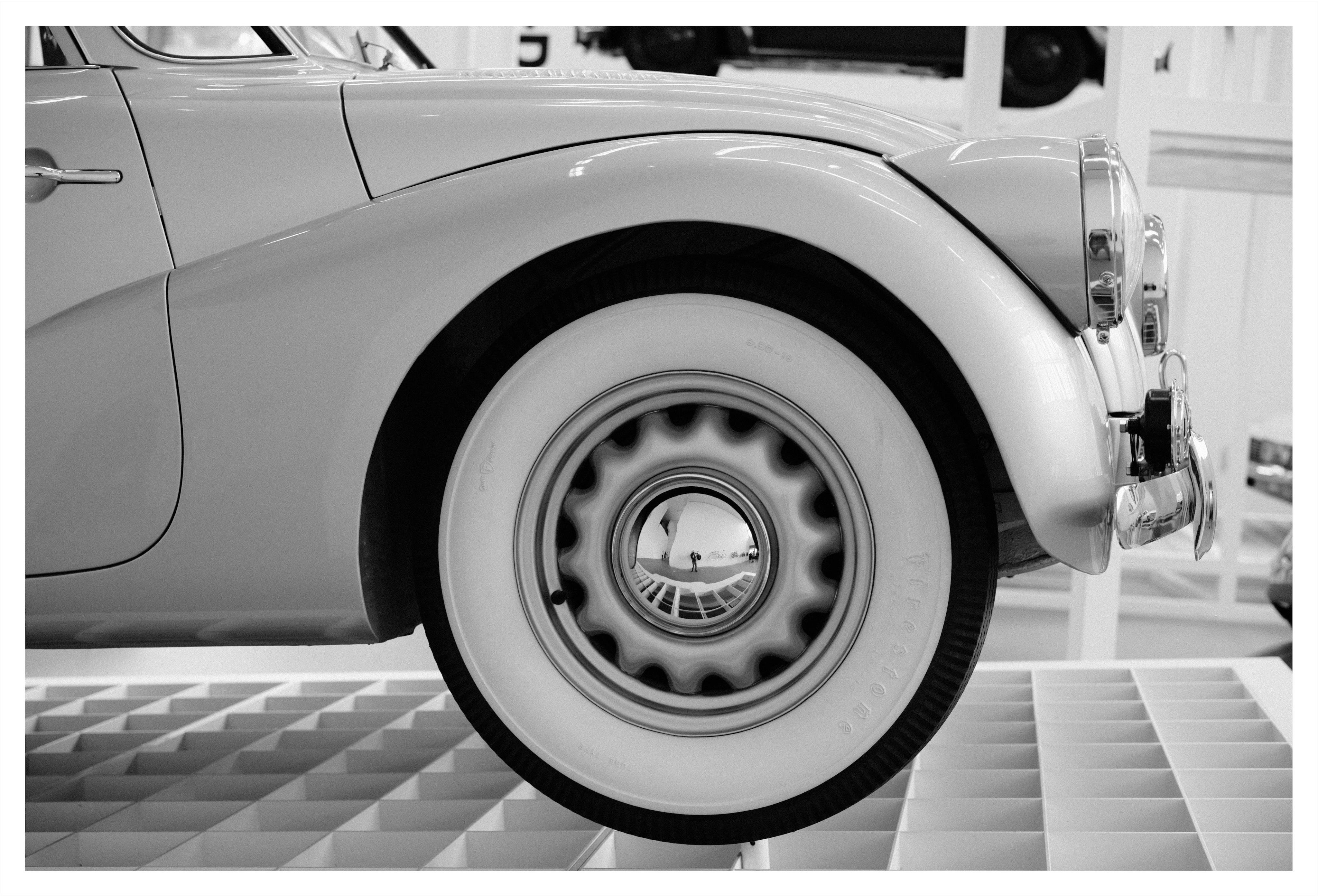 Close-up side view of a vintage car’s front fender and whitewall tire, with the headlight and chrome bumper at right and a patterned floor beneath.