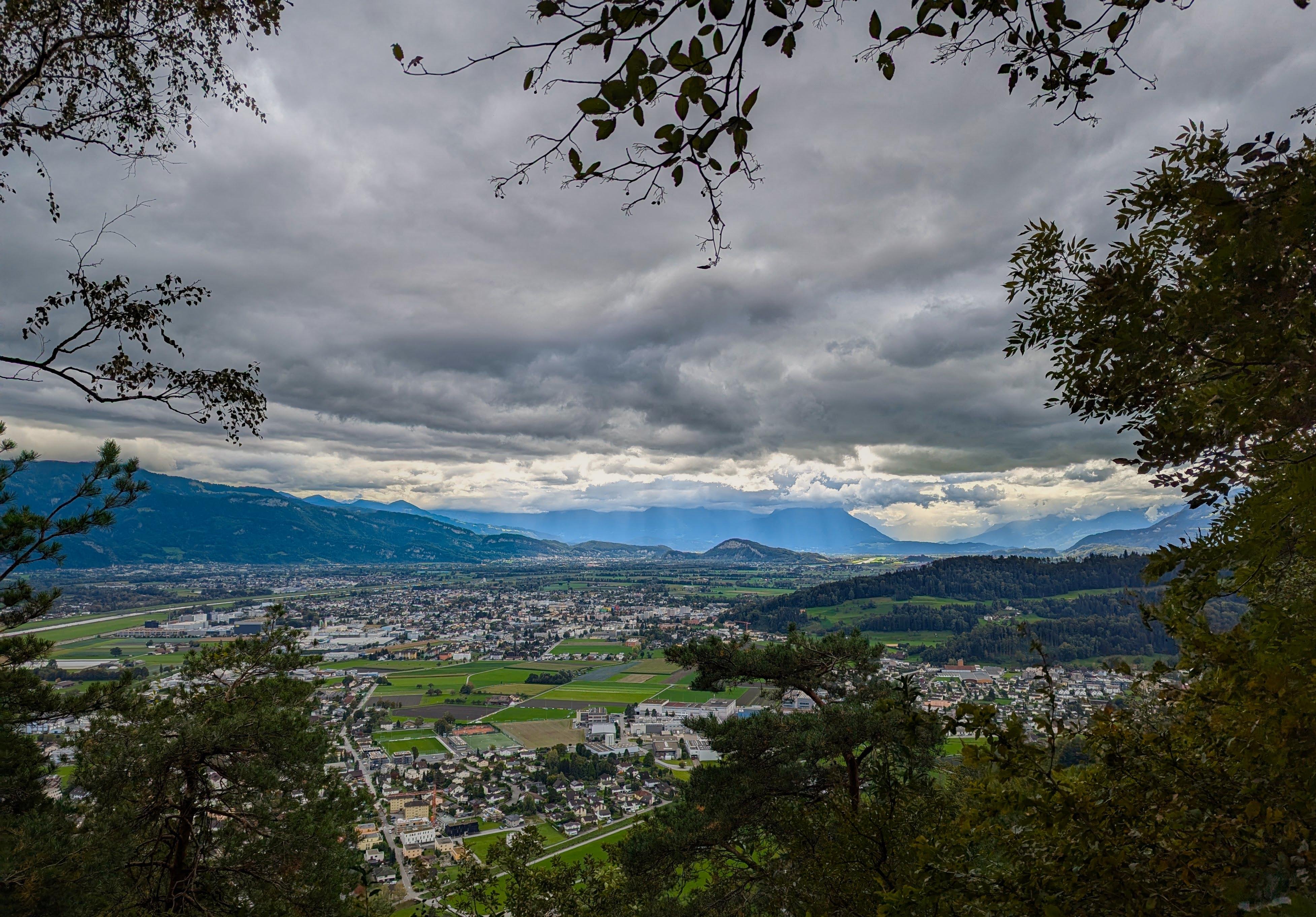 Wide view from a hillside overlooking a valley town with clustered buildings, roads, and patchwork green fields, surrounded by distant mountains under dark, heavy clouds; tree branches and leaves frame the scene at the top and sides.