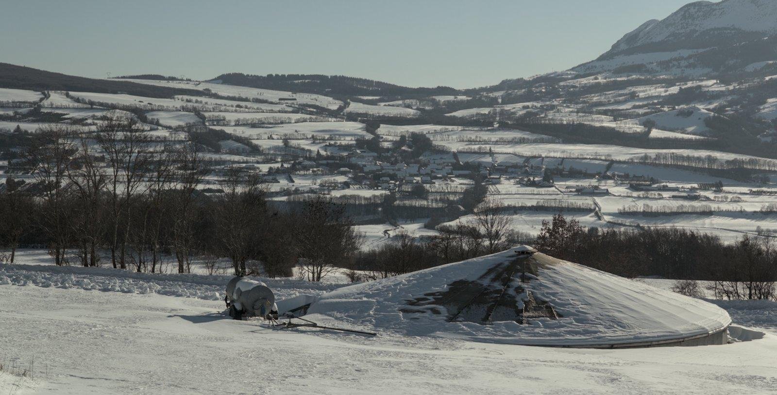Paysage enneigé avec des montagnes en arrière plan, quelques pins, et une structure circulaire avec un toit un peu pointu qui dépasse de la neige et qui fait penser à une soucoupe volante