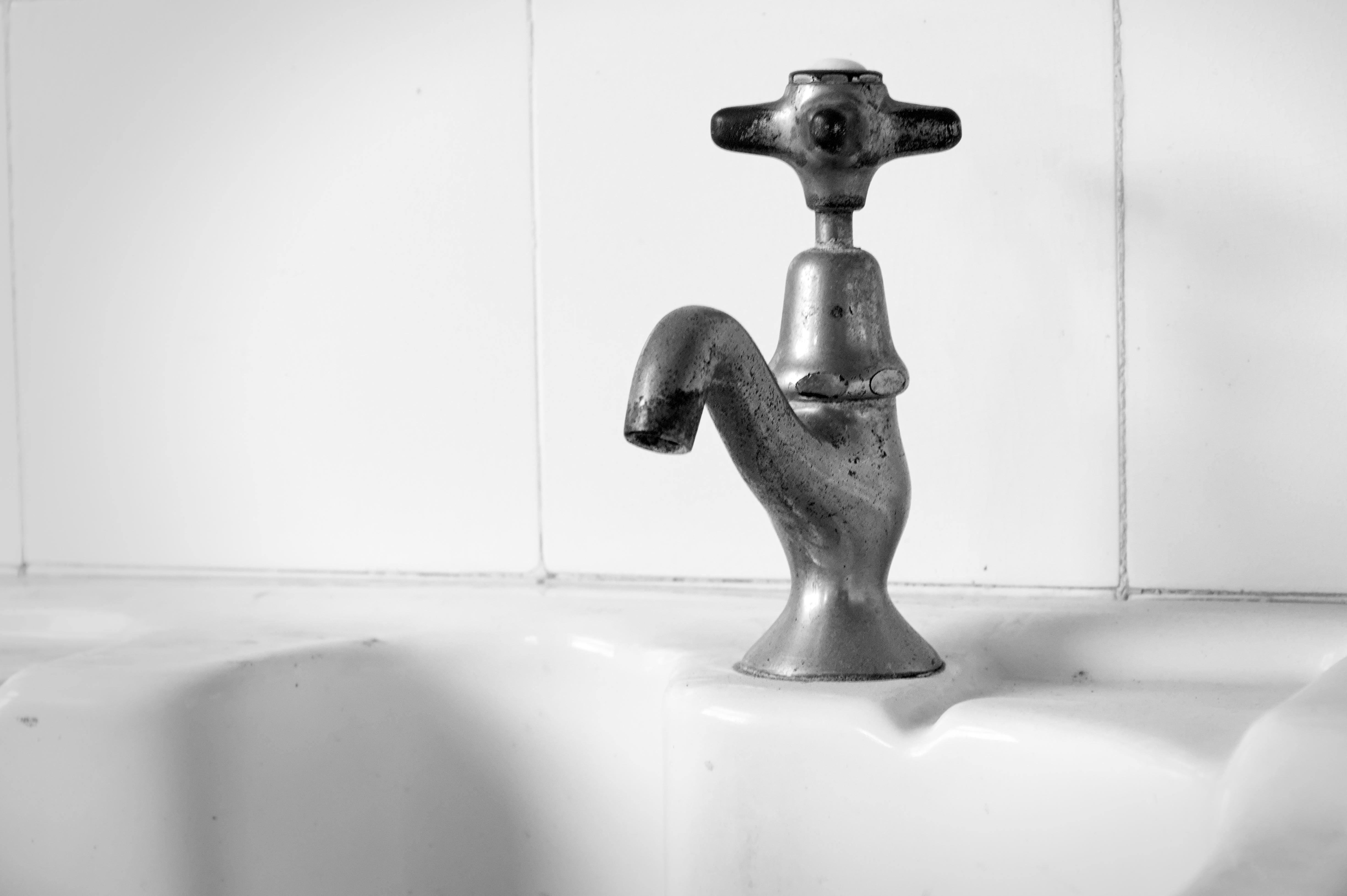 Metallic faucet with a curved spout and a cross-shaped handle stands on a white ceramic sink. Background features plain white tiled wall. The faucet appears slightly tarnished, with visible wear. 
###
Ein metallischer Wasserhahn mit gebogenem Auslauf und kreuzförmigem Griff steht auf einem weißen Keramikwaschbecken. Im Hintergrund befindet sich eine schlichte weiß geflieste Wand. Der Wasserhahn wirkt leicht angelaufen und weist sichtbare Gebrauchsspuren auf. 