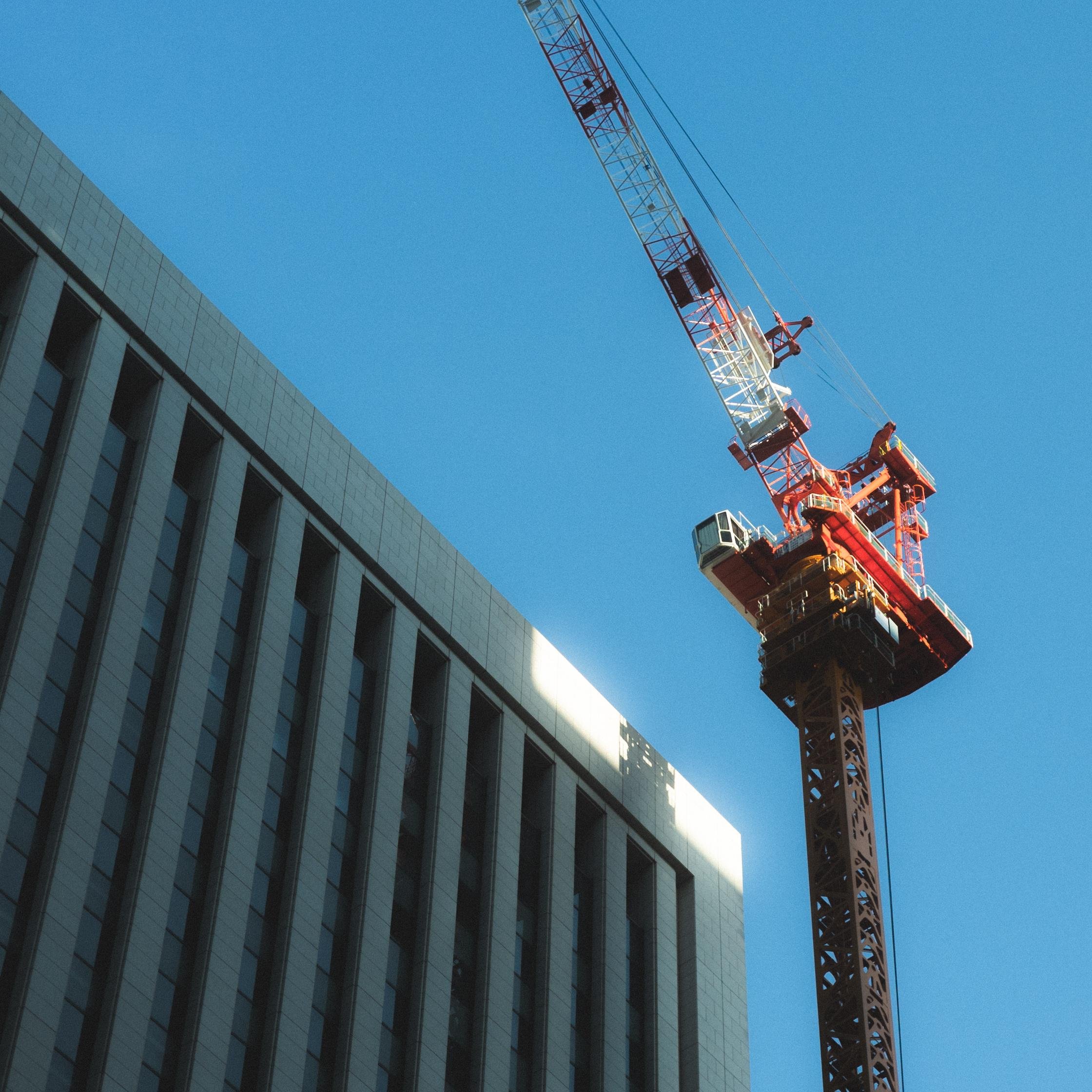 Construction crane near a modern office building with vertical windows, set against a clear blue sky. The sun casts a shadow of the crane onto the building facade.