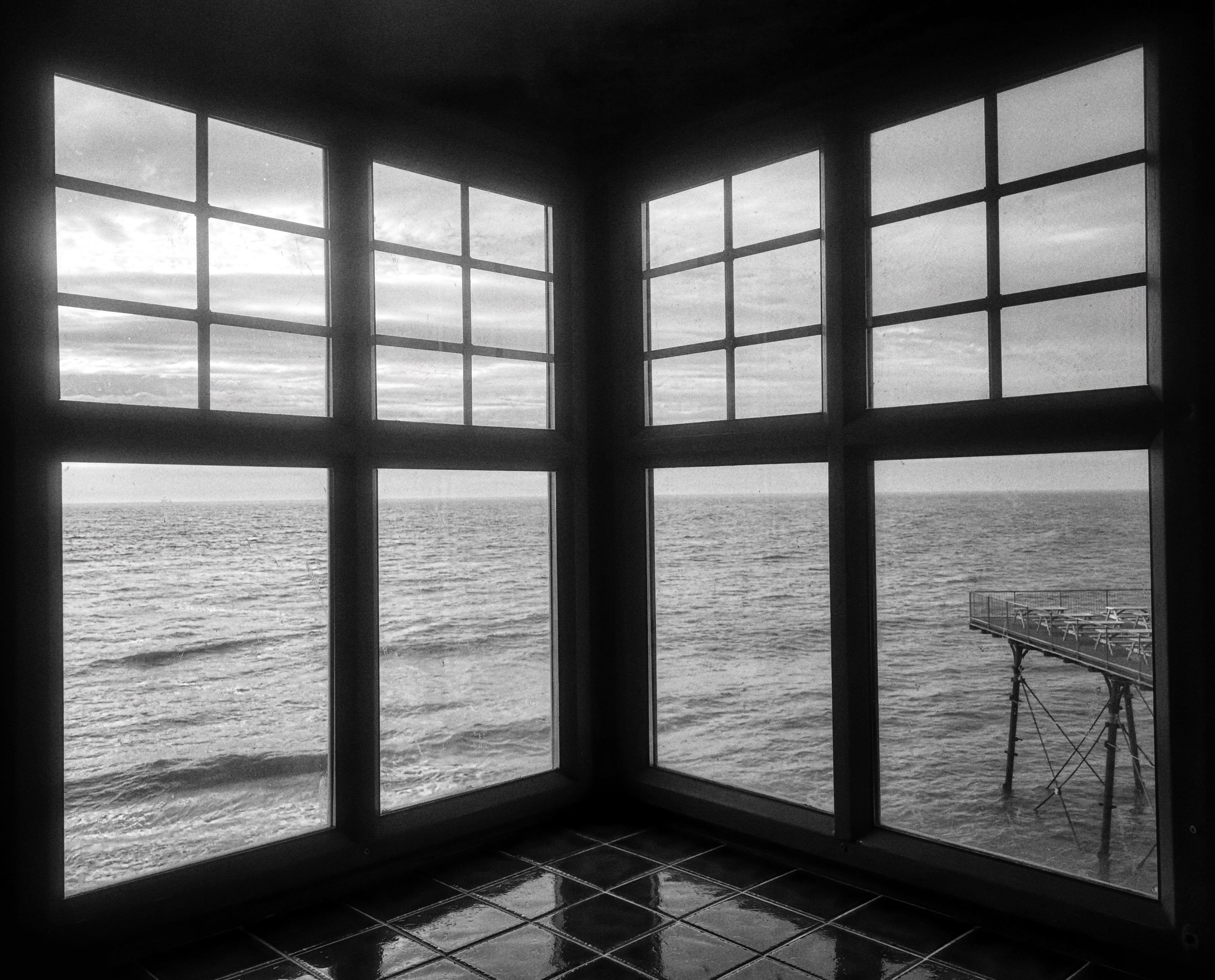 Black-and-white view from inside a the gent's toilets of the Aberystwyth pier, looking out through two large multi-pane windows onto the Irish Sea and cloudy horizon; a tiled floor reflects the window light, and a pier extends into the water on the right.
