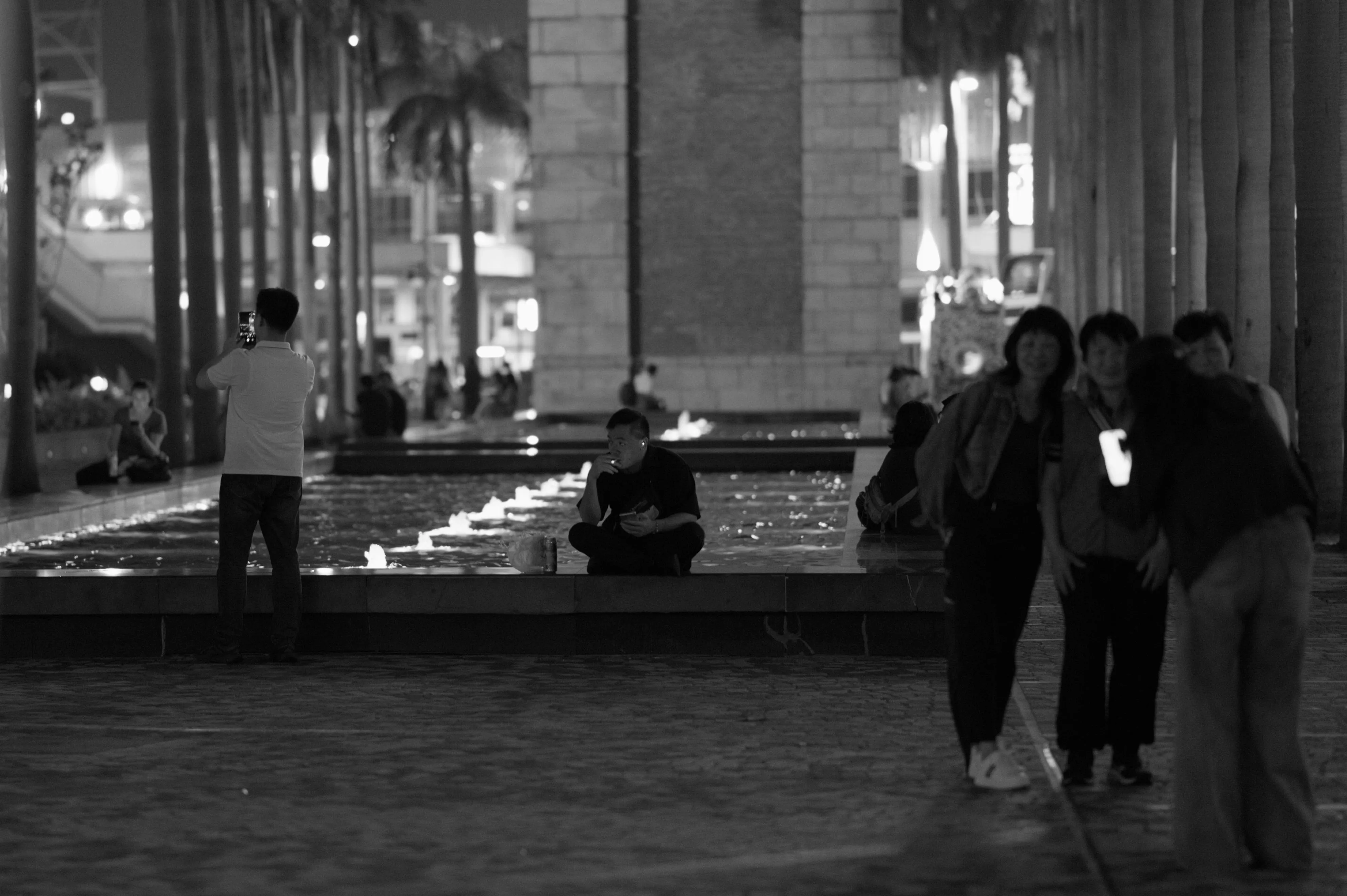 Monochrome nighttime photograph of people in front of a long rectangular fountain basin. On the left side, a man is looking away to the other side of the basin and taking a photograph using his phone. In the middle, a man is sitting cross-legged facing the camera, smoking a cigarette, with earbuds in his ears, a canned drink by his side. The man's earbud and cigarette end constitute two dots of light that align with the lights of the fountains in the basin. The cross-legged man appears to be looking at the man on the left. On the right side in the foreground blur, a group of women are posing together for a group shot with another woman photographing them using a smartphone. In the background, palm trees and the base of a stone tower can be seen.