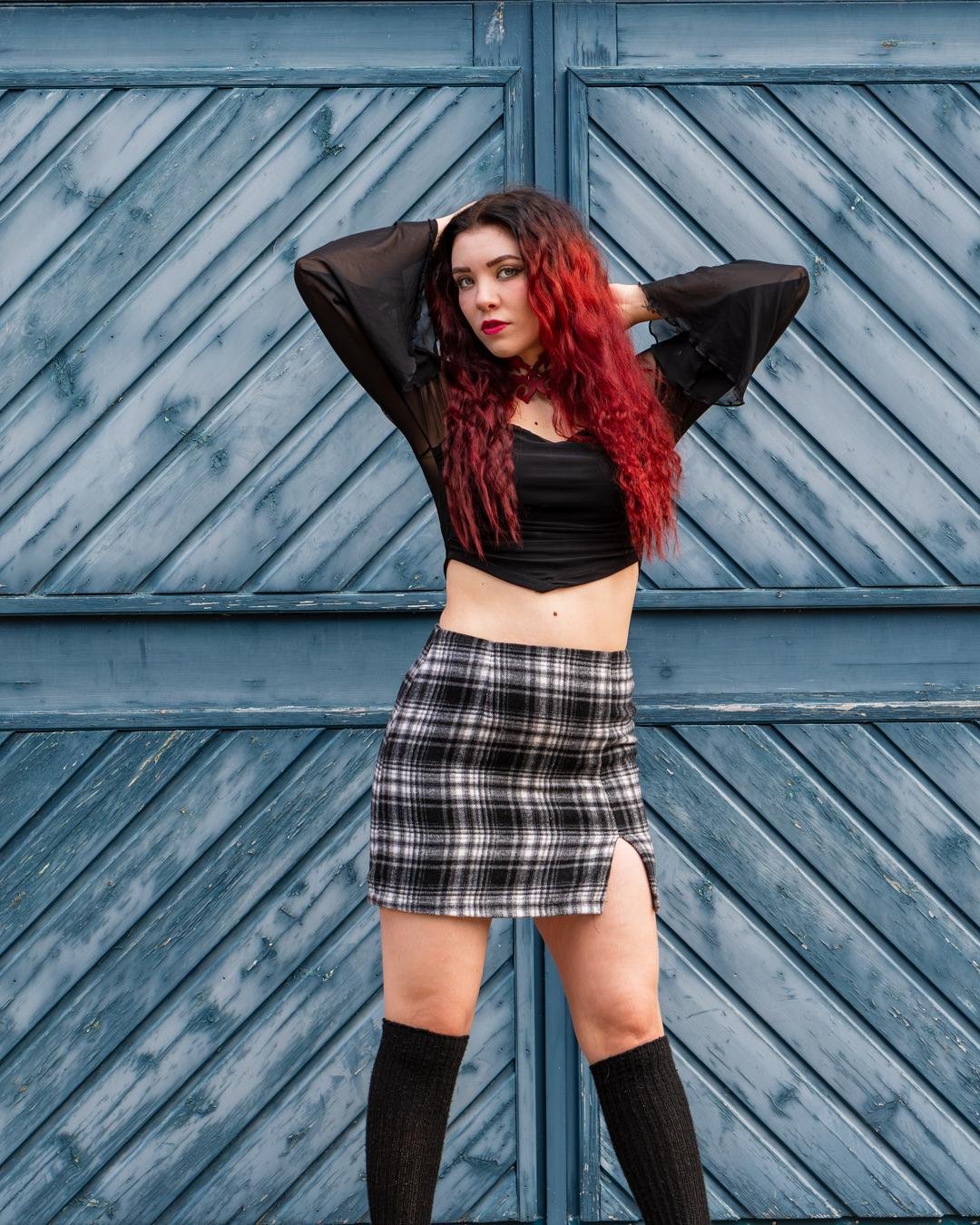 The image shows a person with red hair standing in front of a textured blue wooden door. They are wearing a black top with see-through sleeves, a plaid skirt, and long black socks. The person has their hands raised behind their head.