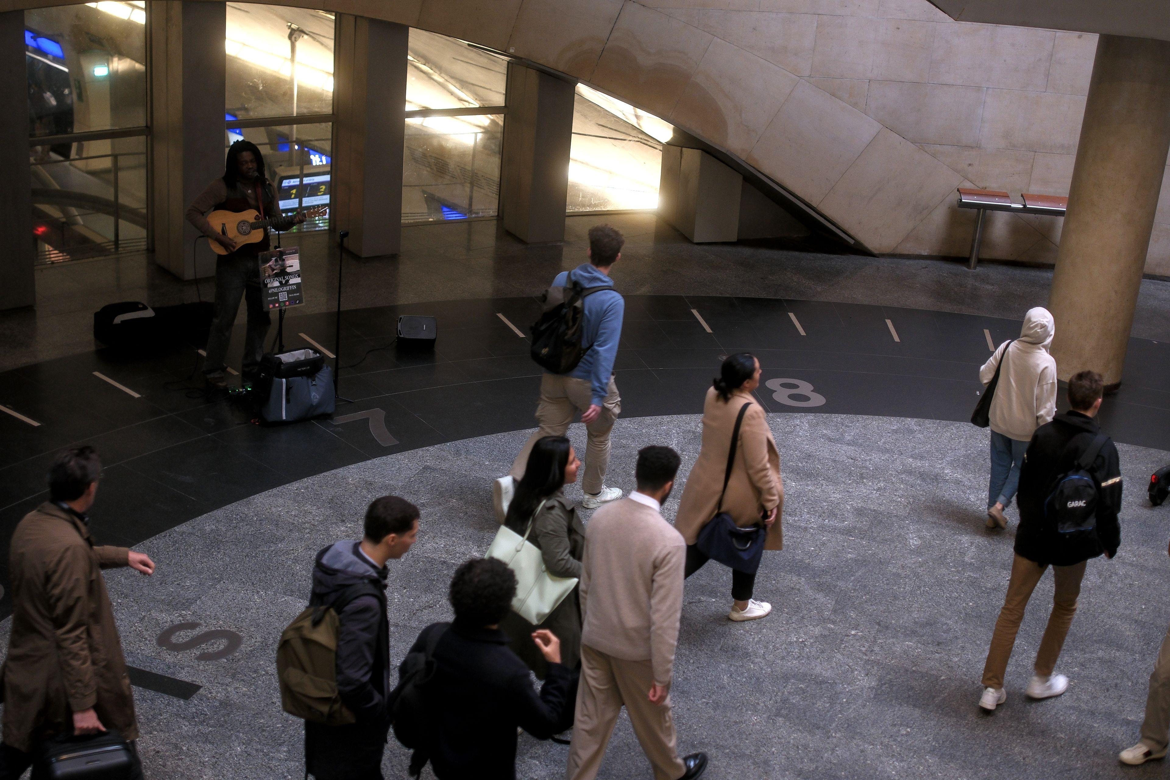 Photo horizontale. Un musicien jouant de la guitare sur une place du métro parisien, des gens passent devant sans lui prêter attention