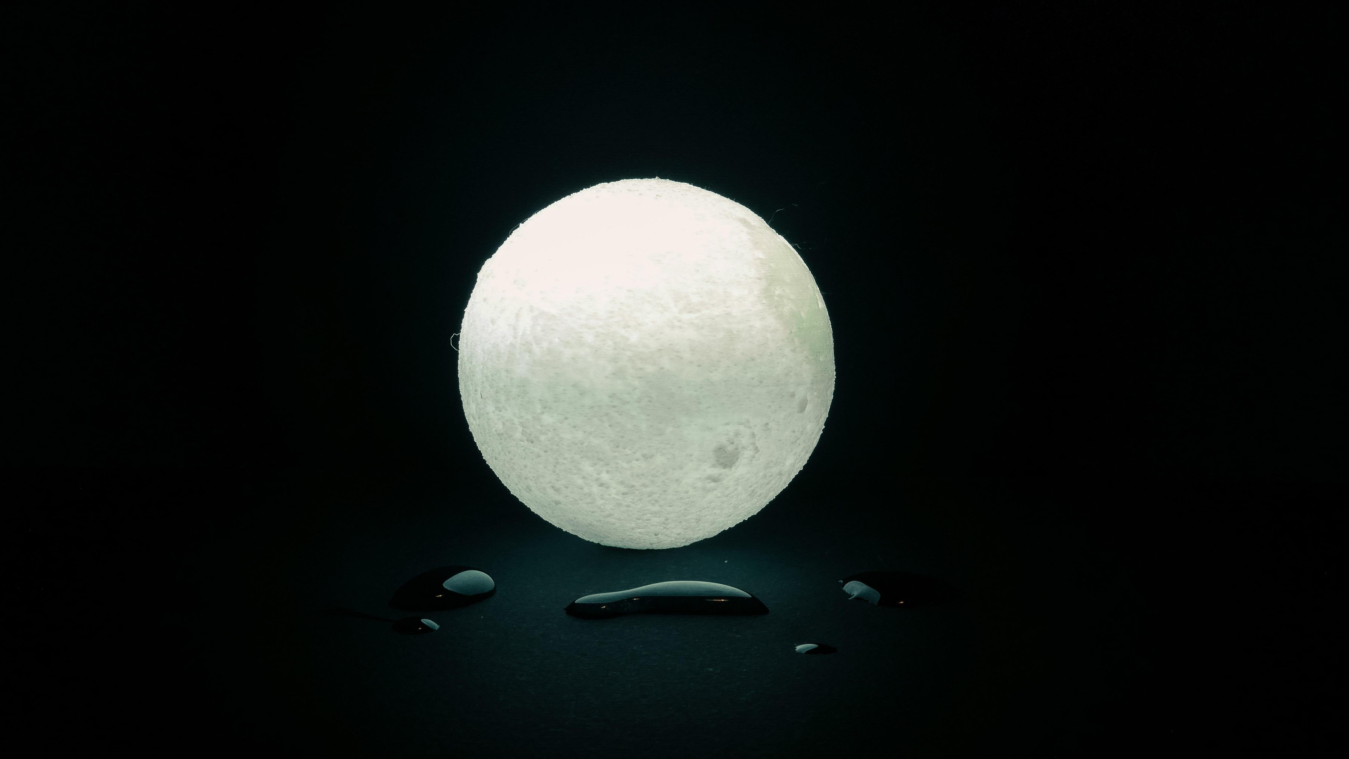 This image features a glowing, spherical object resembling a moon against a dark background. Below the sphere are small water droplets. The setup creates a striking visual contrast.