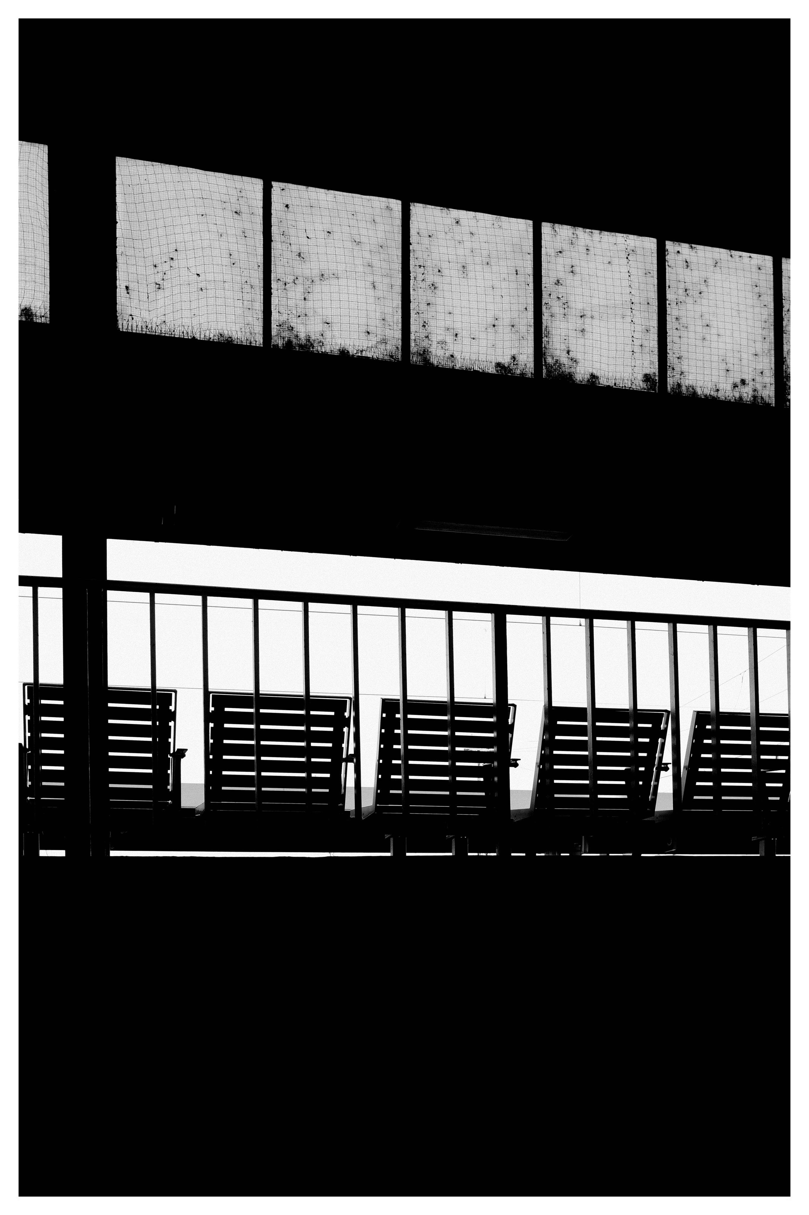 High-contrast black-and-white view of a building facade with a row of rectangular upper windows behind mesh and a lower level of glass panels above silhouetted horizontal slatted shutters.