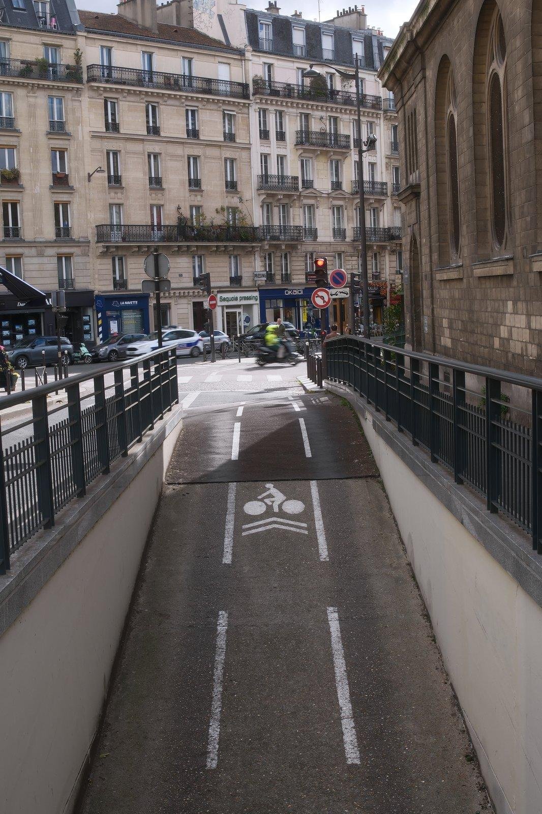 Photo format portrait de la sortie d’une voie vélo souterraine, menant à un rayon de soleil, dans lequel un scooter monté par une personne avec un gilet jaune fluo passe, flou. Le fond est occupé par des immeubles parisiens