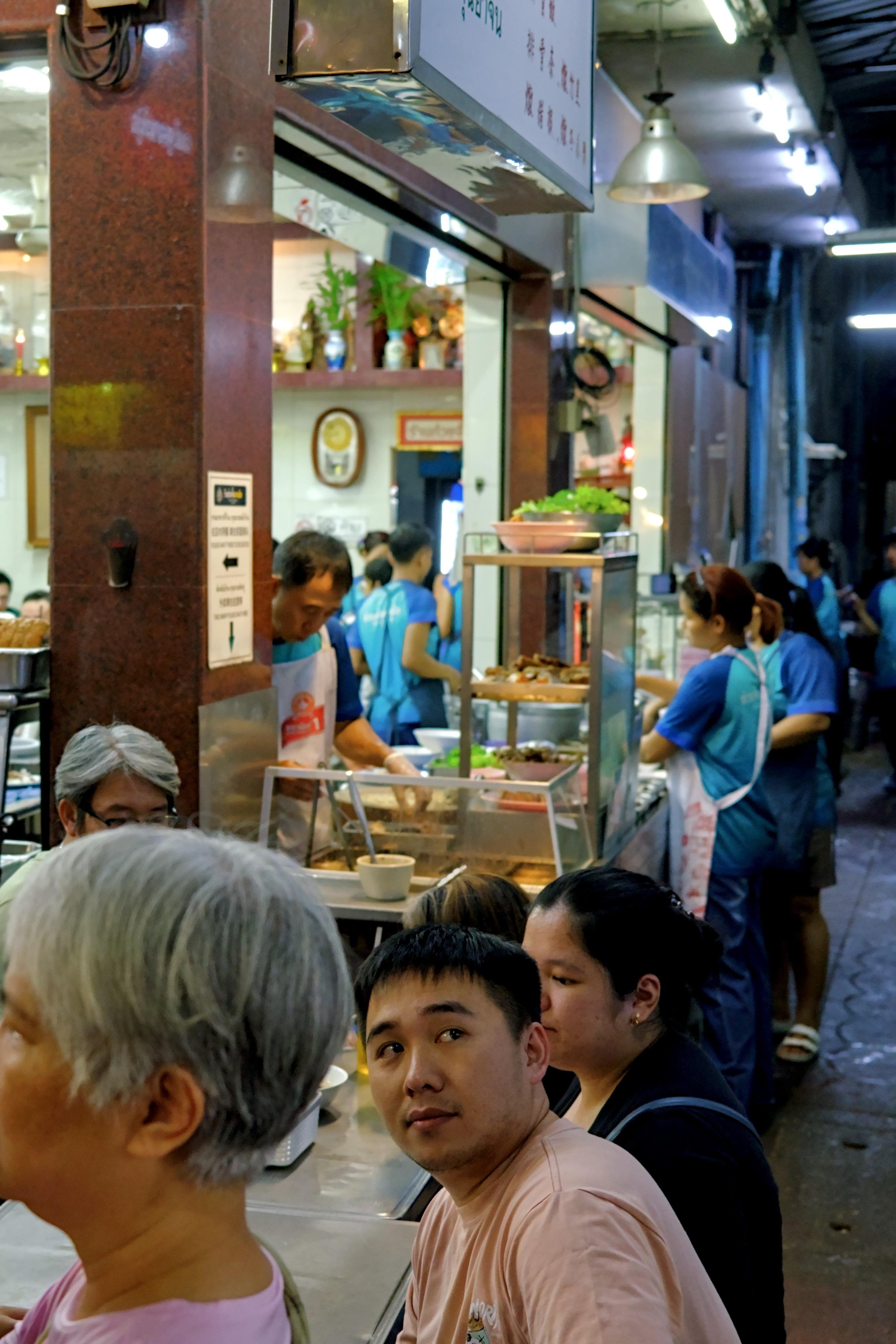 Portrait shot of a street food stall with staff in blue shirts and aprons in the background. In the foreground, a few customers are sat at metal tables. One of them, a young man, the only person in focus, looks back over his shoulder, a strong side-eye as he attempts to look at something behind the camera.