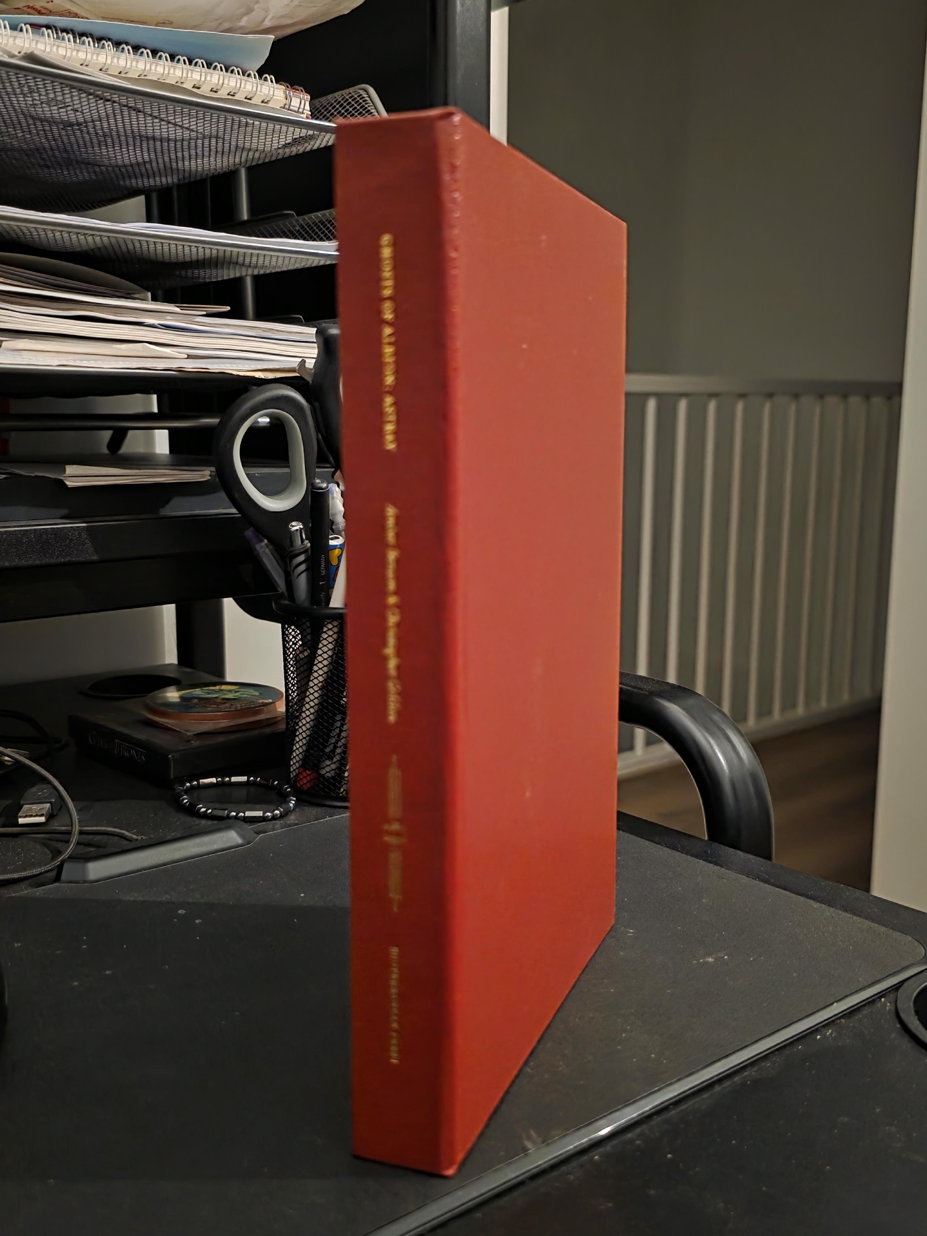 The image shows a side view of a special edition of the novel "Ghosts of Albion: Astray" by Amber Benson and Christopher Golden. The book is housed in a sleek, matching brick-red slipcase that stands vertically on a dark desk. On the spine of the slipcase, the title and authors' names are elegantly stamped in gold foil, giving it a premium, collectible feel.
This specific release is a limited edition from Subterranean Press, restricted to only 750 numbered copies. Astray is the second novel in the Ghosts of Albion series, continuing the adventures of Tamara and William Swift as they protect Victorian London from supernatural threats. This edition is highly sought after by collectors for its high-quality production and its connection to the rich urban fantasy world originally created as a BBC animated series. The background shows a casual home office setting, emphasizing the book as a prized part of a personal collection.