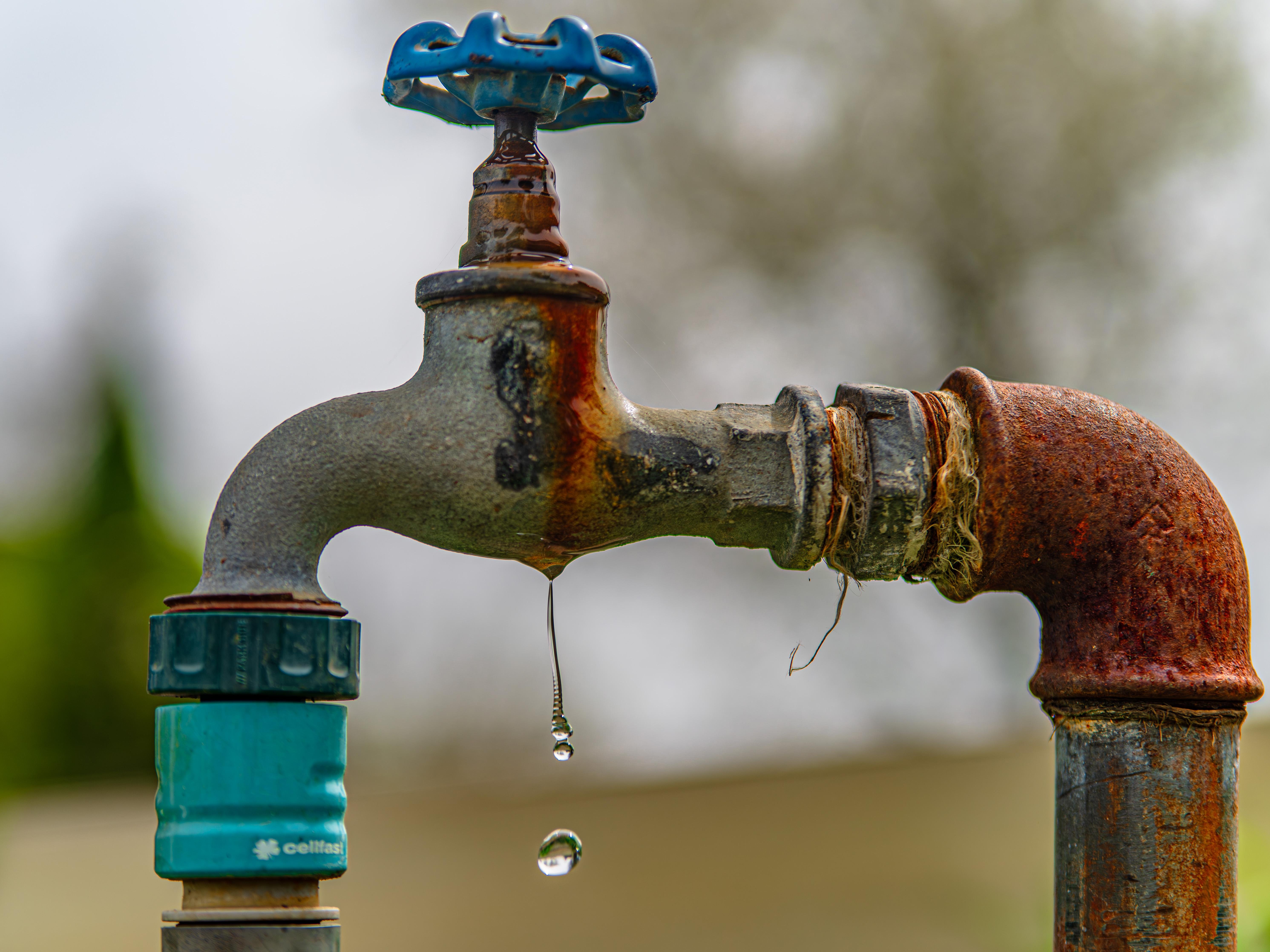 Close-up of a rusty outdoor water faucet with two droplets hanging from the spout. The metal surface shows signs of wear and oxidation. The handle on top is blue, and a green connector is attached below. The background is blurred, suggesting an outdoor setting.
