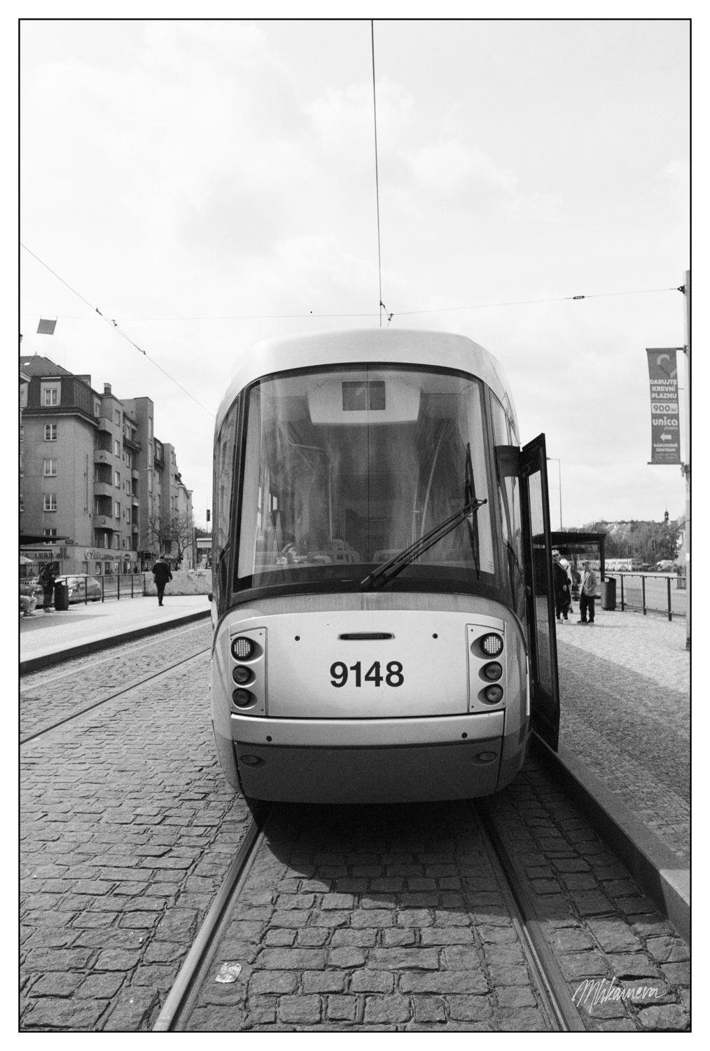 A monochrome photo of a rear end of a modern tram standing in a stop with clouds reflecting in the round rearscreen window