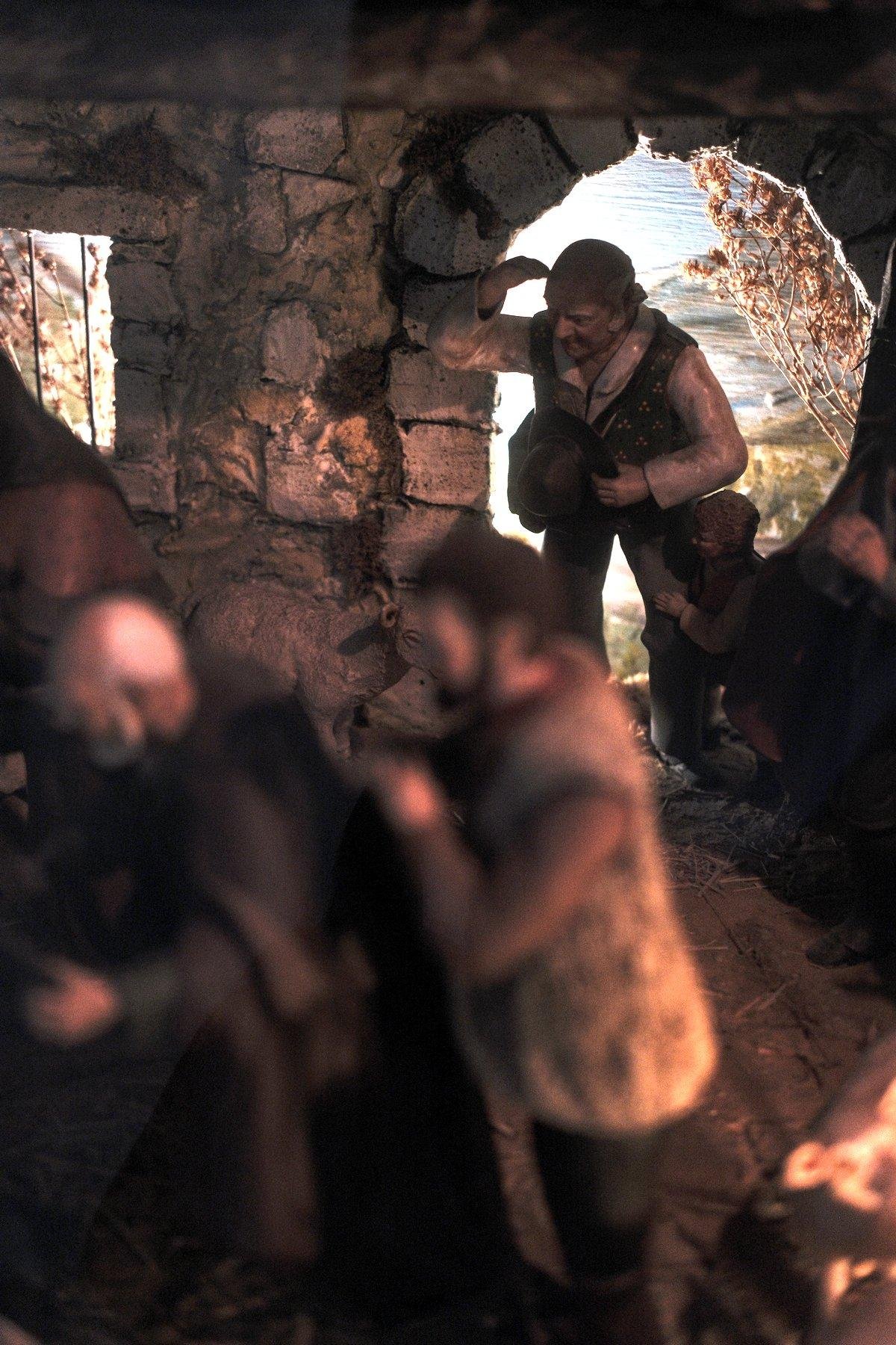 Photo verticale. Une scène en santon montrant un homme dans l’embrasure de la porte d’un bâtiment, portant sa main en visière, son enfant est accroché à sa jambe de timidité. Au premier plan, flou, des hommes semble se grouper pour voir un truc