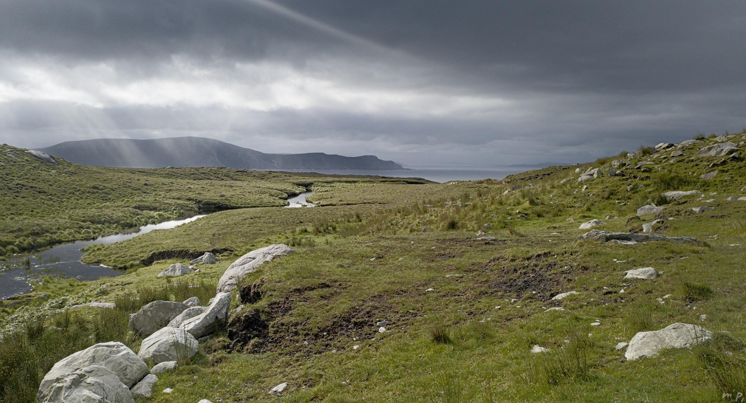 🇬🇧 Horizontal color photo of an Irish landscape in the morning. In the foreground, white rocks strewn on a hilly green moor, in the background the coast where we can see a little of sea, the distant relief on the other side of a bay, all under a charged sky where rays pierce oblique sun.

🇫🇷 Photo couleur horizontale d'un paysage d'Irlande au matin. Au premier plan, des rochers blancs semés sur une lande verte valonnée, au second plan la côte où on distingue un peu de mer, le relief lointain de l'autre côté d'une baie, le tout sous un ciel chargé où percent des rayons de soleil obliques.