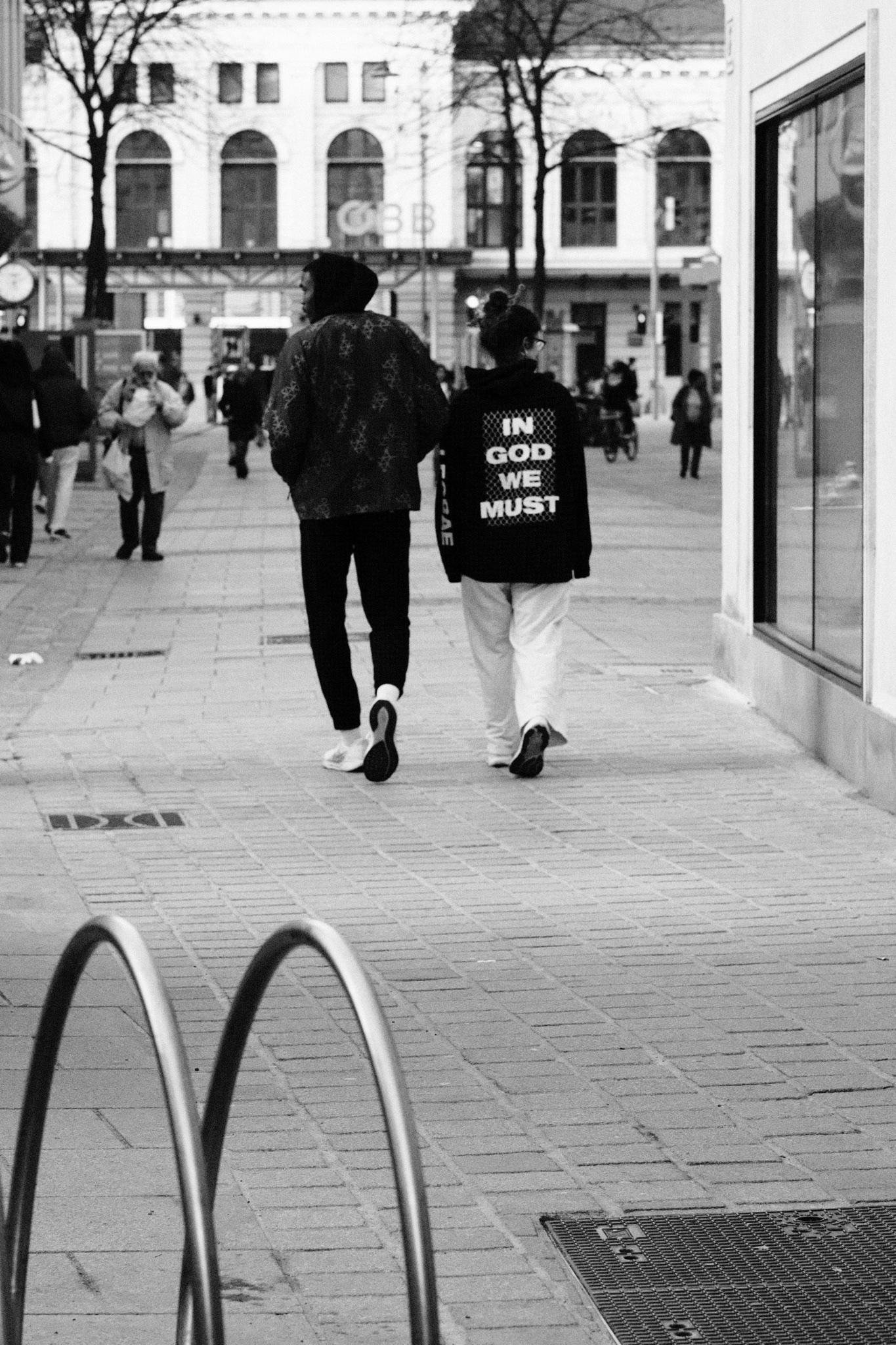 Schwarz-Weiß-Straßenfoto von zwei Personen, die einen Fußgängerzone hinuntergehen; eine trägt eine Jacke mit einem großen Text mit der Aufschrift „IN GOD WE MUST“. Andere Fußgänger und ein mehrstöckiges Gebäude mit gewölbten Fenstern befinden sich im Hintergrund, mit einem Schaufenster auf der rechten Seite und gebogenen Fahrradständerstangen im Vordergrund.