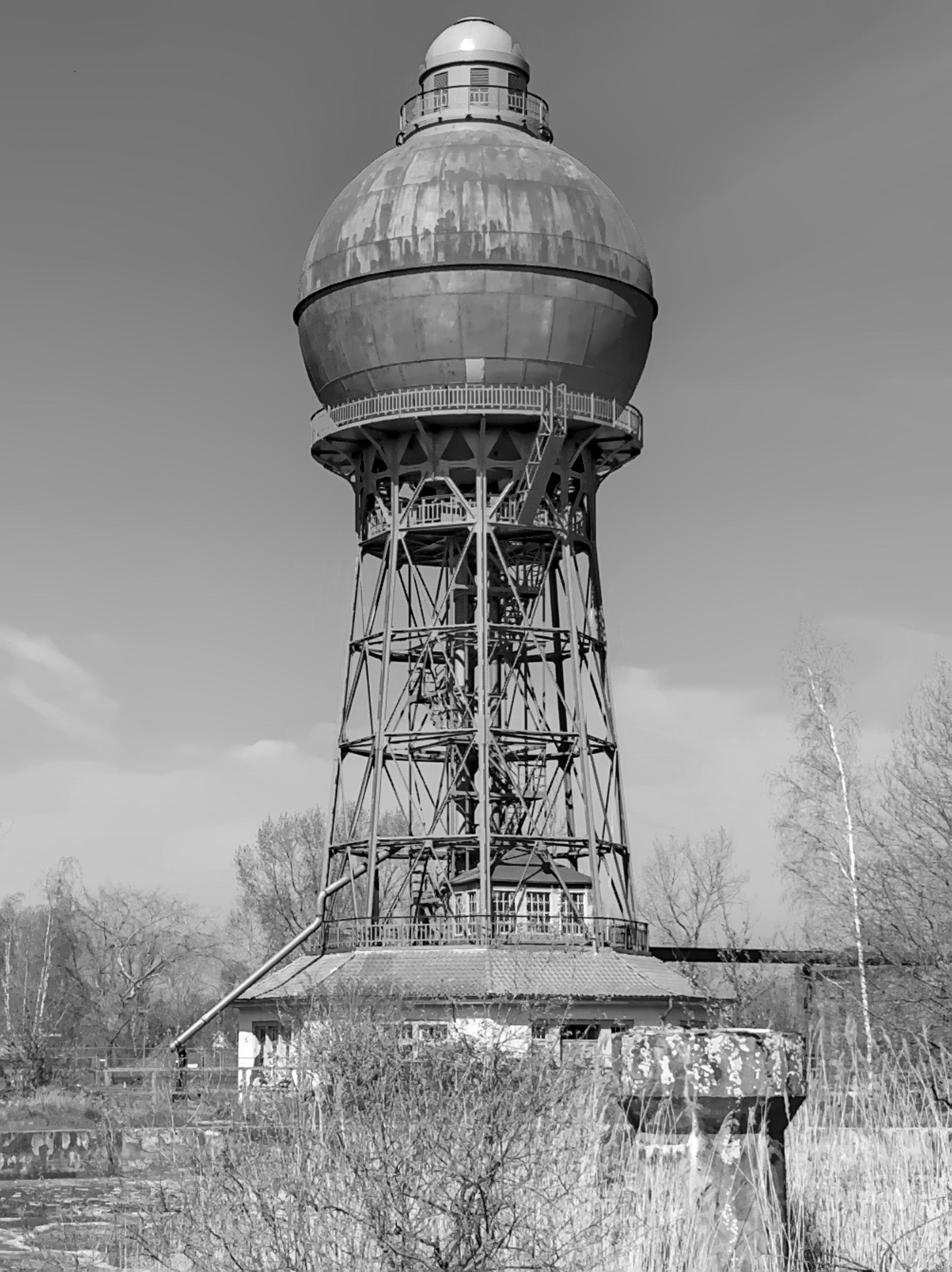 The image shows a large, spherical water tower with a metal support structure. The tower is elevated, suggesting its purpose for storing water and providing pressure to a water supply system. The surrounding area includes some vegetation and appears to be in a somewhat industrial or rural setting.