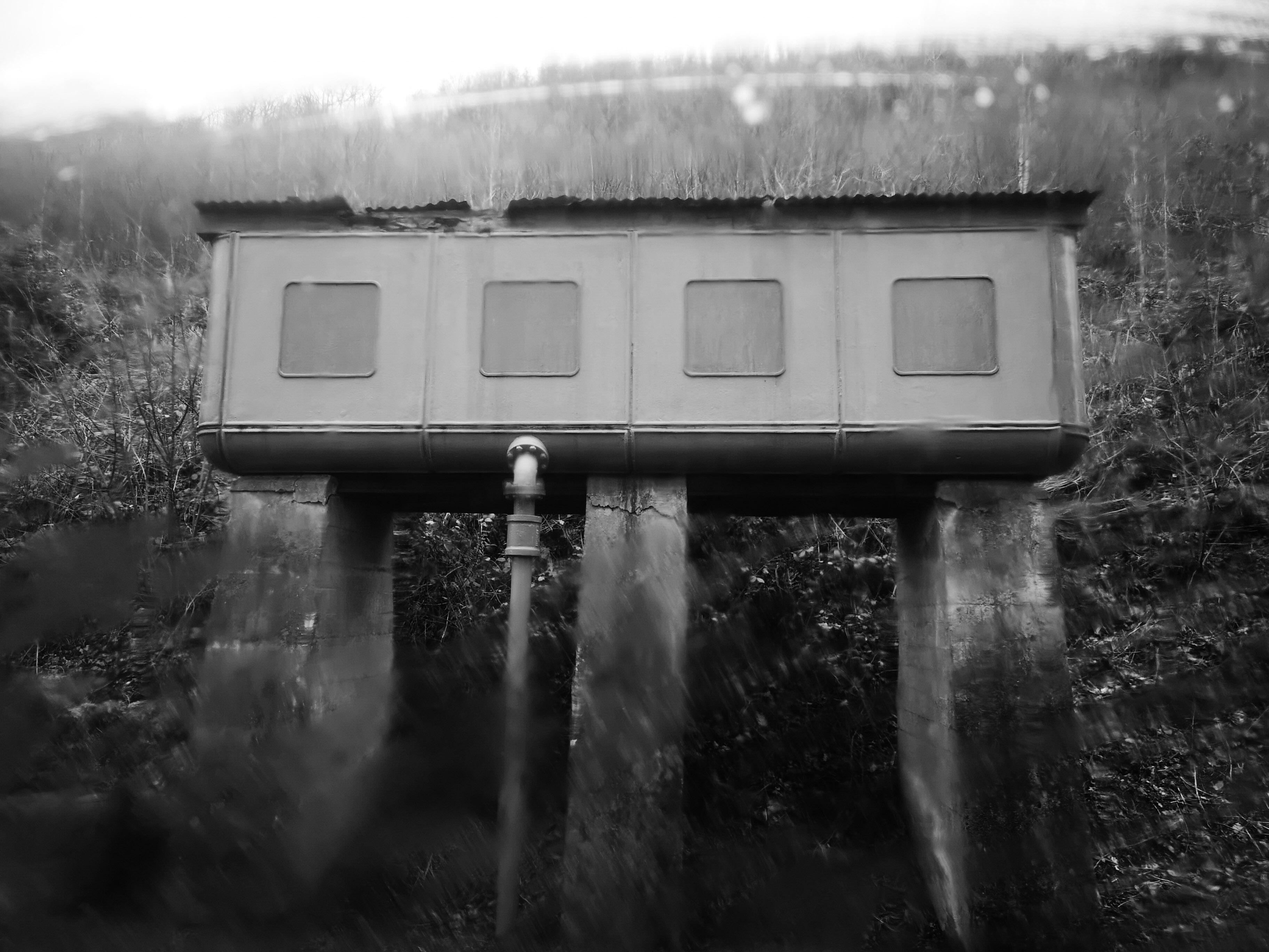 Black-and-white photo of a water tower on tall concrete stilts, with four square window panels along the side and a pipe rising to the underside, set against a brushy hillside. Picture taken from the steamy window of a train.
