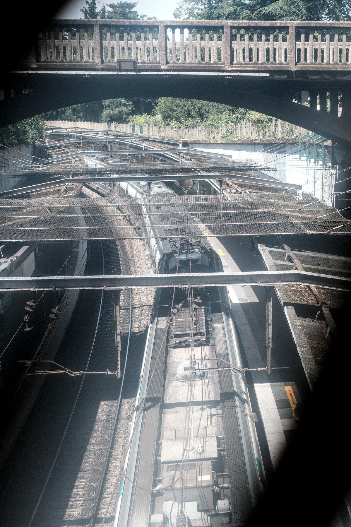Photo verticale. Vue à travers un grillage comme en témoignent des traits noirs au 1er plan dans le coin NW et SE, sur une double voie ferrée en courbe vers la gauche. Sur celle de droite se trouve une rame de RER dans une station à ciel ouvert. Au dessus des poutres métalliques et encore au dessus un pont