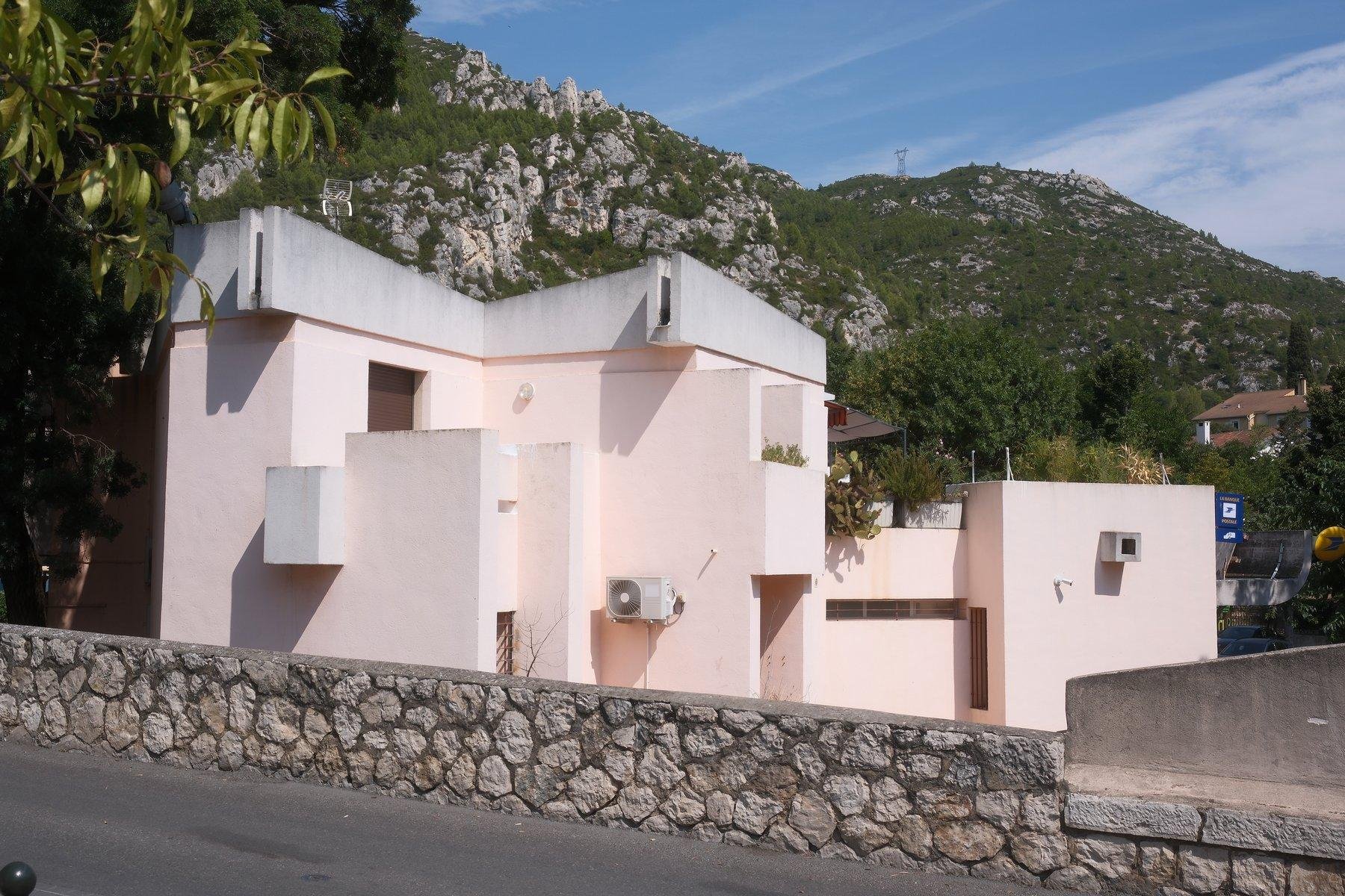 Photo horizontale. Un bâtiment à l’architecture très cubique et majoritairement rose pastel, avec le sommet gris clair