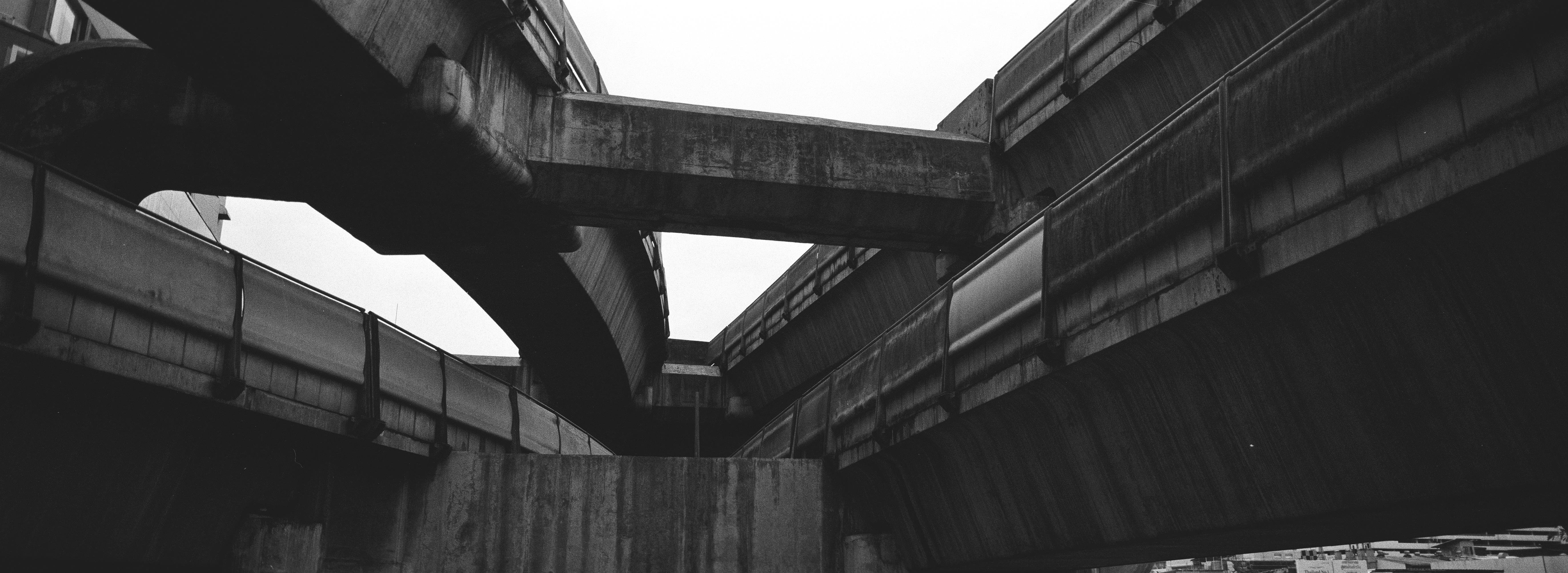 Black and white panoramic photo of the sky train's intersecting concrete overpasses in a complex, angular arrangement. Four bridges are joining on two levels, connected by a concrete beam against a white sky background