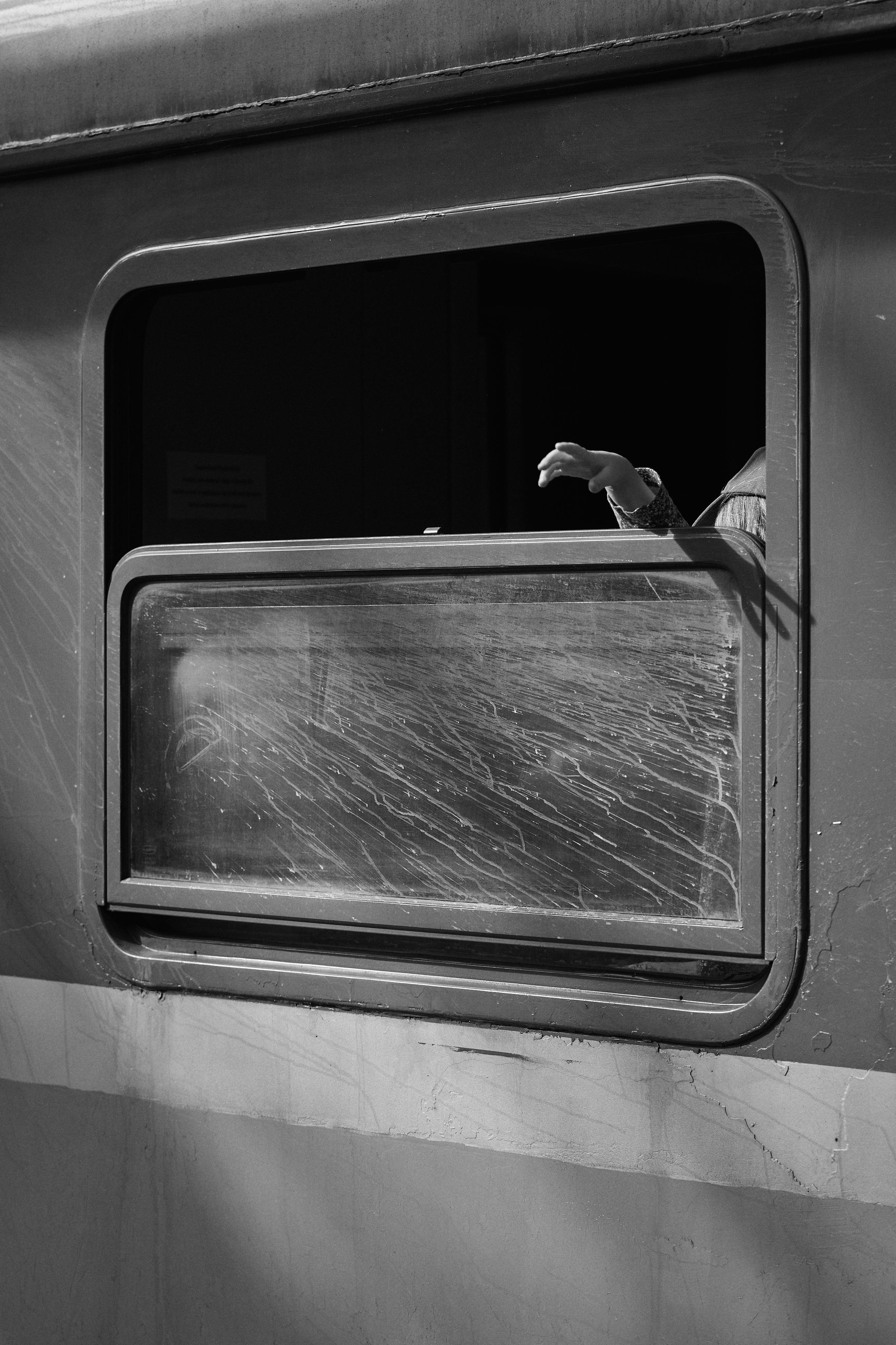 Black and white image of a partially open train window with a scratched surface. An arm extends out of the window, with the hand slightly raised. The exterior of the train shows signs of wear. Shadows are visible on the train's surface.