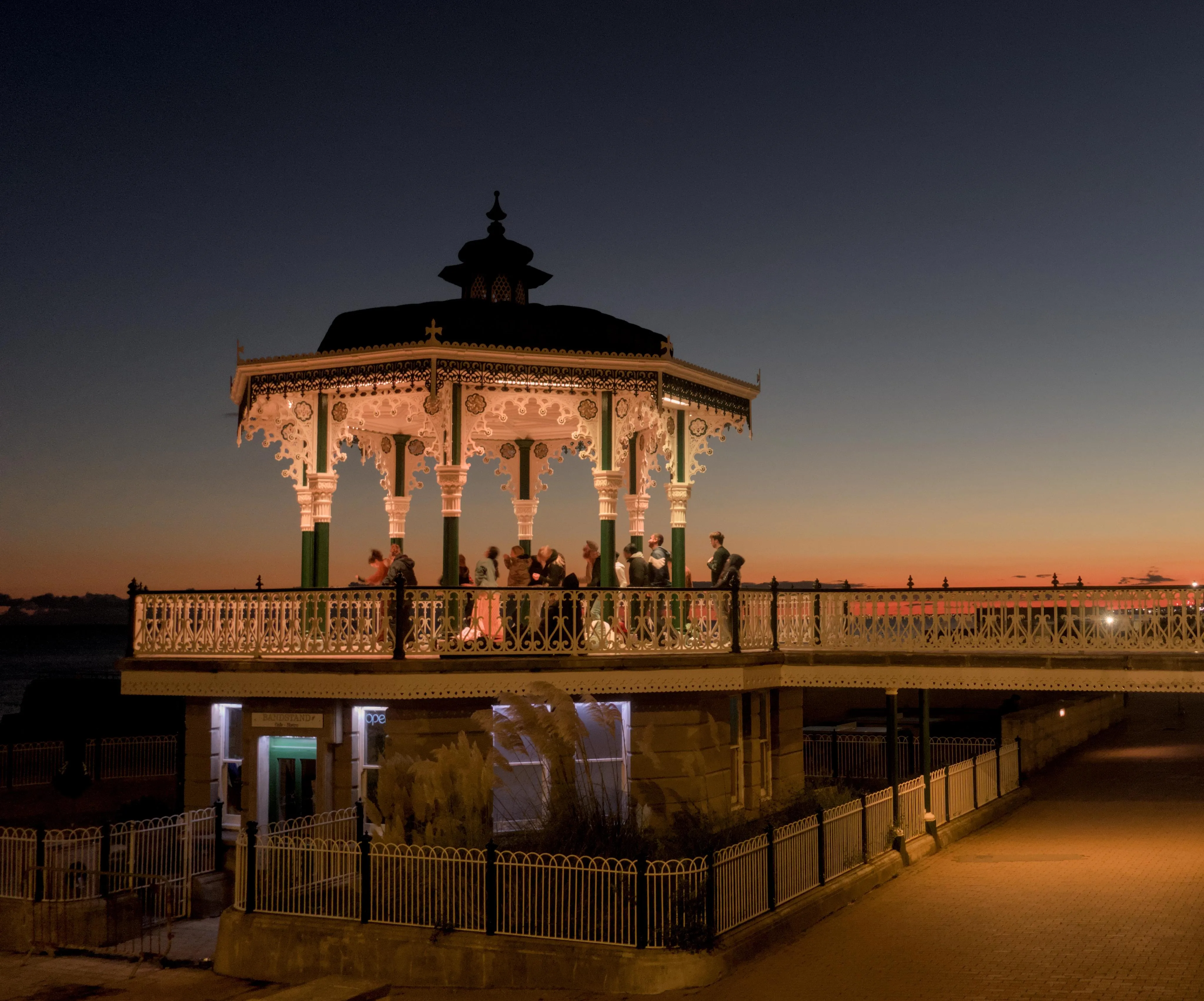 Photograph of dance group training at sunset in a gazebo by a seafront promenade. The sunset sky is a gradient from deep blue at the top to yellow to red near the horizon. The dancers are blurry from motion blur while the gazebo and other static elements of the picture are detailed and neat.