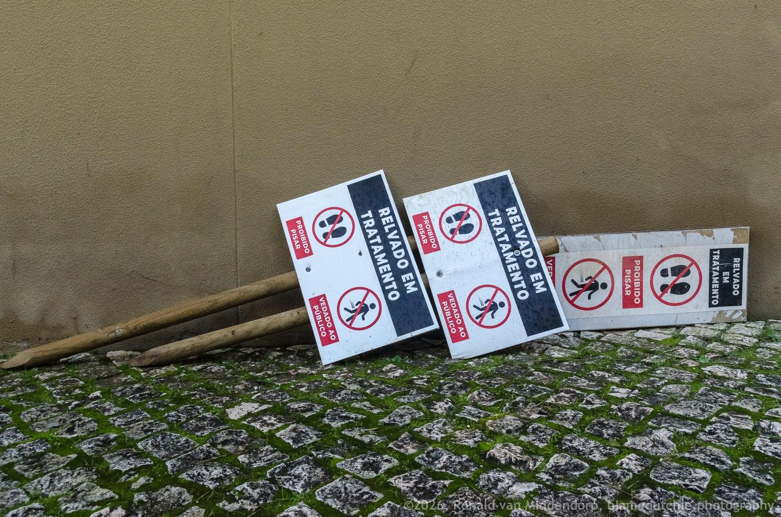 Three warning signs reading “RELVADO EM TRATAMENTO” with red prohibition icons lie on mossy cobblestones against a beige wall.