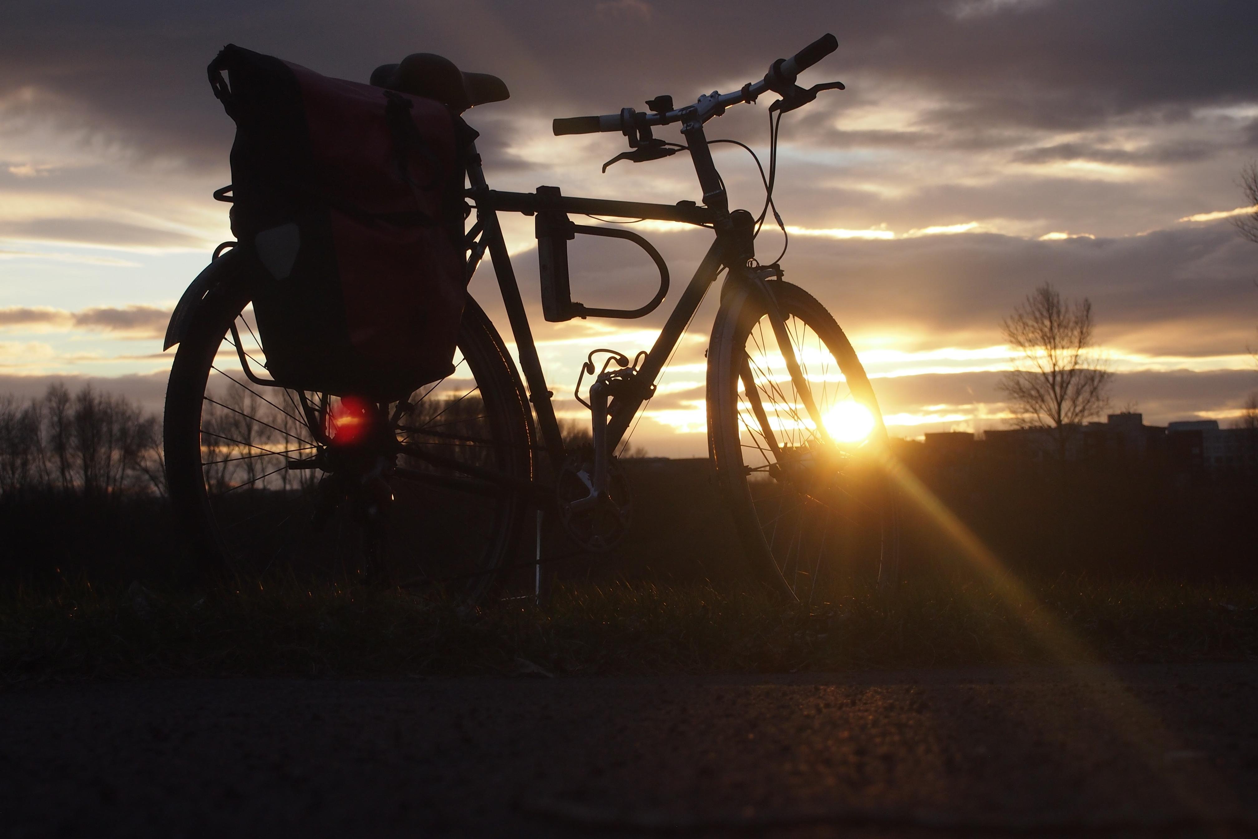 Foto im Querformat. Die Kamera befindet sich knapp über dem Boden.
Im Vordergrund steht ein Fahrrad mit einer roten Fahrradtasche. Durch das Vorderrad scheint die untergehende Sonne, Sonnenstrahlen reichen bis in die rechte untere Ecke. Im Hintergrund befinden sich blätterlose Bäume und Häusersiloutten. Der Himmel ist mit dunklen Wolken bedeckt, ihre Kanten sind durch das Sonnenlicht hervorgehoben.